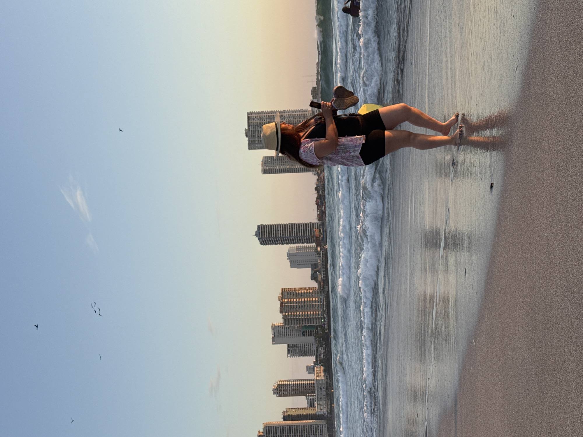 A vertical medium-range shot depicts a woman standing at the edge of a beach, her feet in the water. She is fair-skinned with dark hair, wearing a straw hat, a printed short-sleeve shirt, and black shorts. She holds a cell phone and appears to be taking a selfie. Beside her hangs a pair of brown sandals, and a yellow, oblong object is tucked at her waist. In the background, high-rise buildings line the horizon, suggesting a coastal city, and birds fly across the clear sky. The beach's wet sand reflects the sky and buildings, and waves gently touch the shoreline.