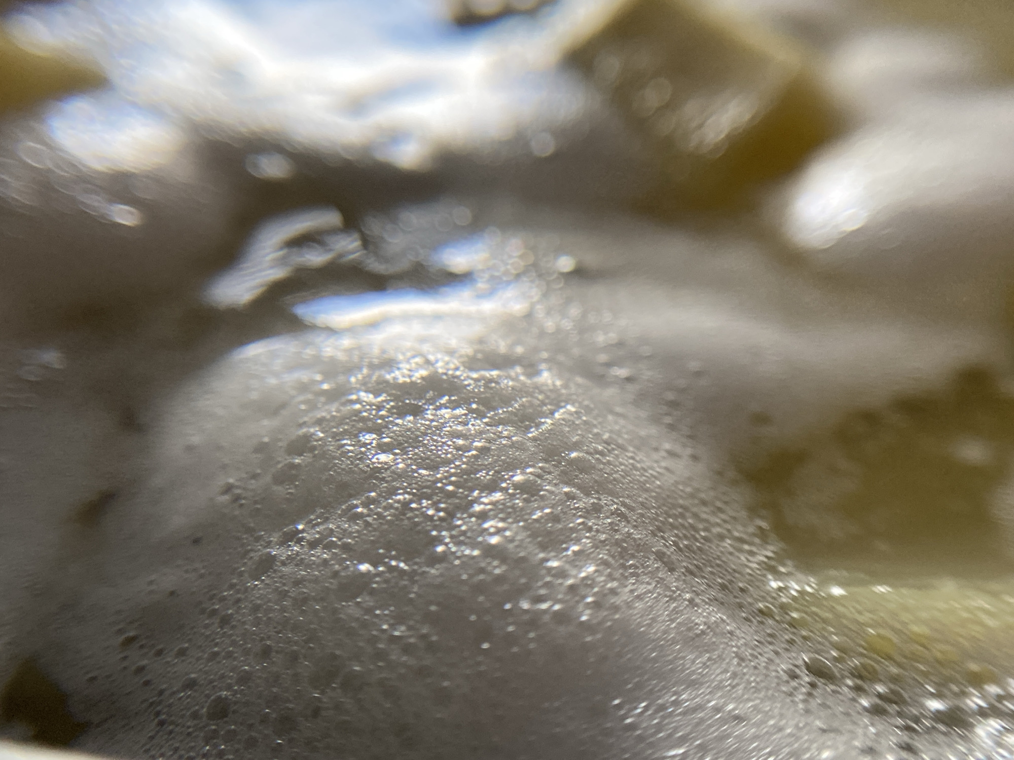 The image is a close-up, high-angle shot of a foamy, bubbly surface, likely liquid being heated or agitated. The foam is dense and appears to be a light, off-white color. Within the foam, small, shimmering bubbles are visible, catching the light and creating a textured effect. In the background, out of focus, there are hints of a yellowish-brown liquid and possibly solid objects submerged. The lighting suggests it is daytime, with light reflecting off the bubbles and the surface of the liquid. There are no people or identifiable structures visible. The scene is abstract and focuses on the micro-details of the foamy liquid.
