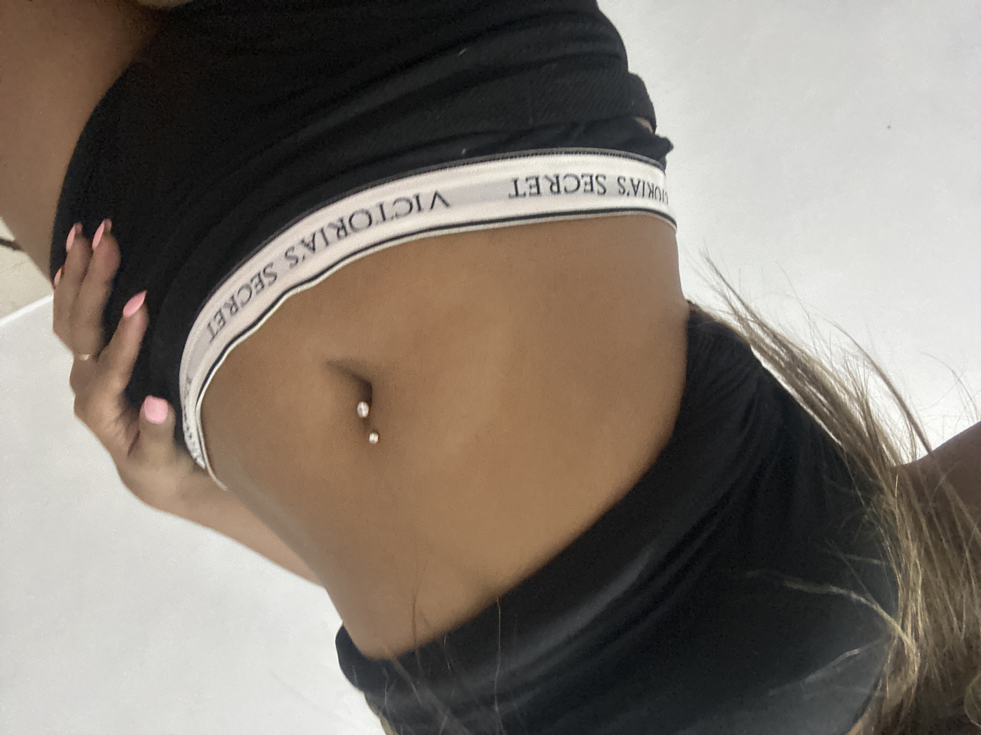 The image shows a close-up of a person's midriff wearing a black crop top and black underwear with "VICTORIA'S SECRET" printed on the waistband. The person has a navel piercing. The background is a plain white wall. The lighting suggests an indoor environment. There is no indication of the location in Sukawati, Indonesia.