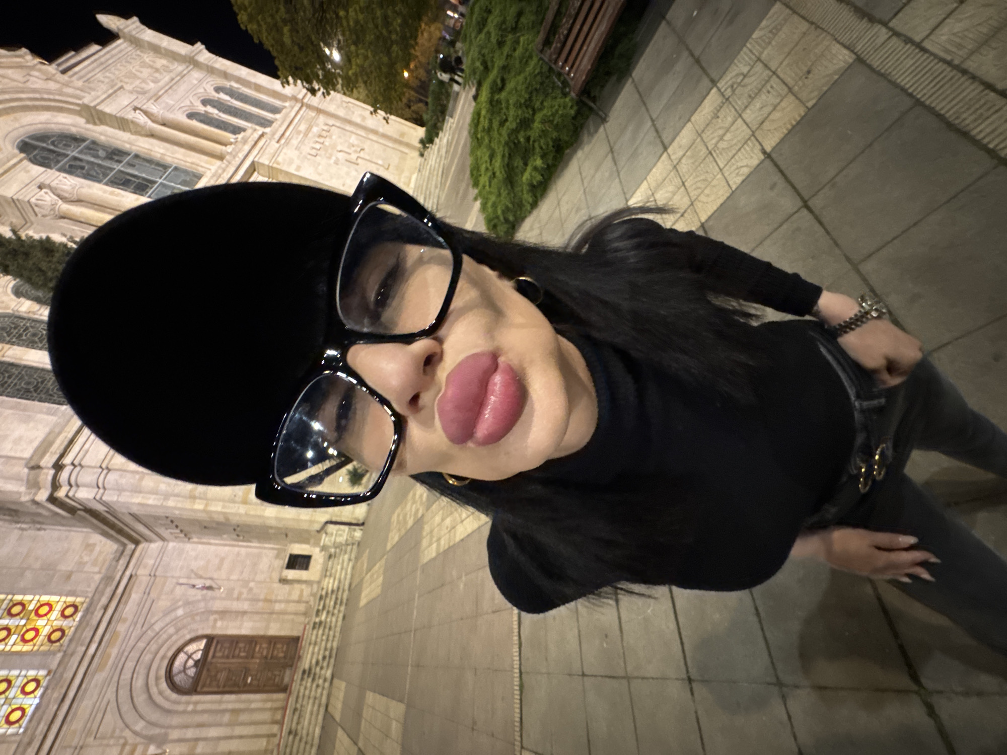A close-up, low-angle selfie captures a person outdoors at night in Varna, Bulgaria. The subject, likely female, faces the camera with full, glossy pink lips in a prominent pout. They wear large, black-framed cat-eye glasses that reflect ambient light, a black wide-brimmed hat, and a black long-sleeved top, possibly a turtleneck. Long dark hair cascades around the shoulders, and gold hoop earrings are visible. A multi-strand dark beaded bracelet or watch adorns the left wrist, and long, manicured nails are visible on both hands.
The background features an ornate, light-colored stone building, likely a church or cathedral, characterized by classical architectural details such as arched windows, elaborate carvings, and visible stained glass with red and yellow circular and semi-circular patterns. The scene is illuminated by artificial lights, casting shadows and highlighting the building's facade against a dark night sky. The ground is paved with patterned tiles, indicative of a city sidewalk or plaza. In the mid-ground, there is dense green foliage and a wooden park bench, suggesting a landscaped area adjacent to the building. No legible text is visible.