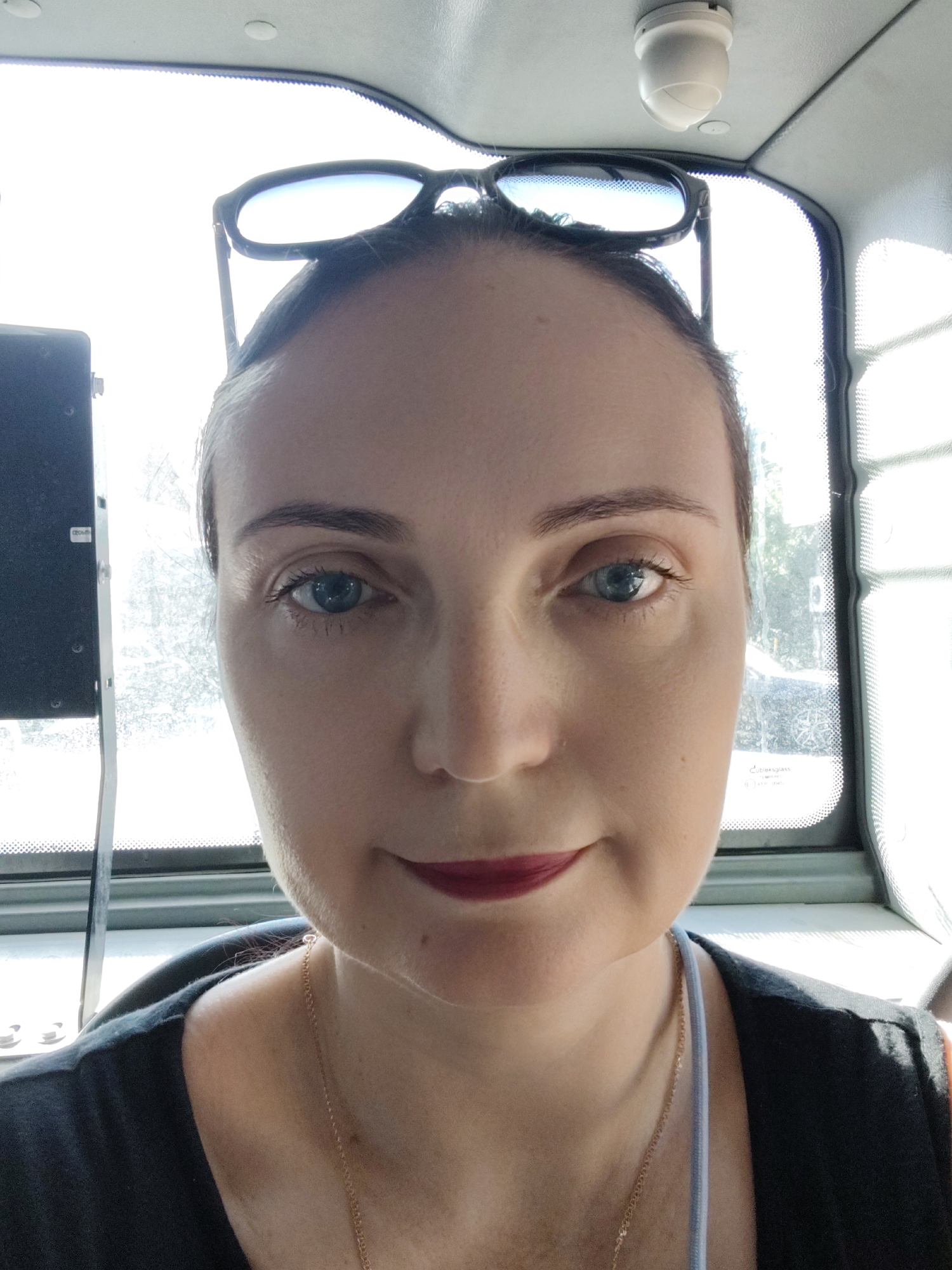 A woman is taking a selfie in a vehicle. She has dark hair pulled back, and is wearing sunglasses on top of her head. She is wearing dark clothing, and a gold necklace. Her lips are painted with dark red lipstick. She appears to be looking directly at the camera. The background shows a window with some greenery visible outside, and a white ceiling with a light fixture. The scene appears to be inside a bus or a similar vehicle. There is no discernible indication of the specific location within Sochi, Russia.