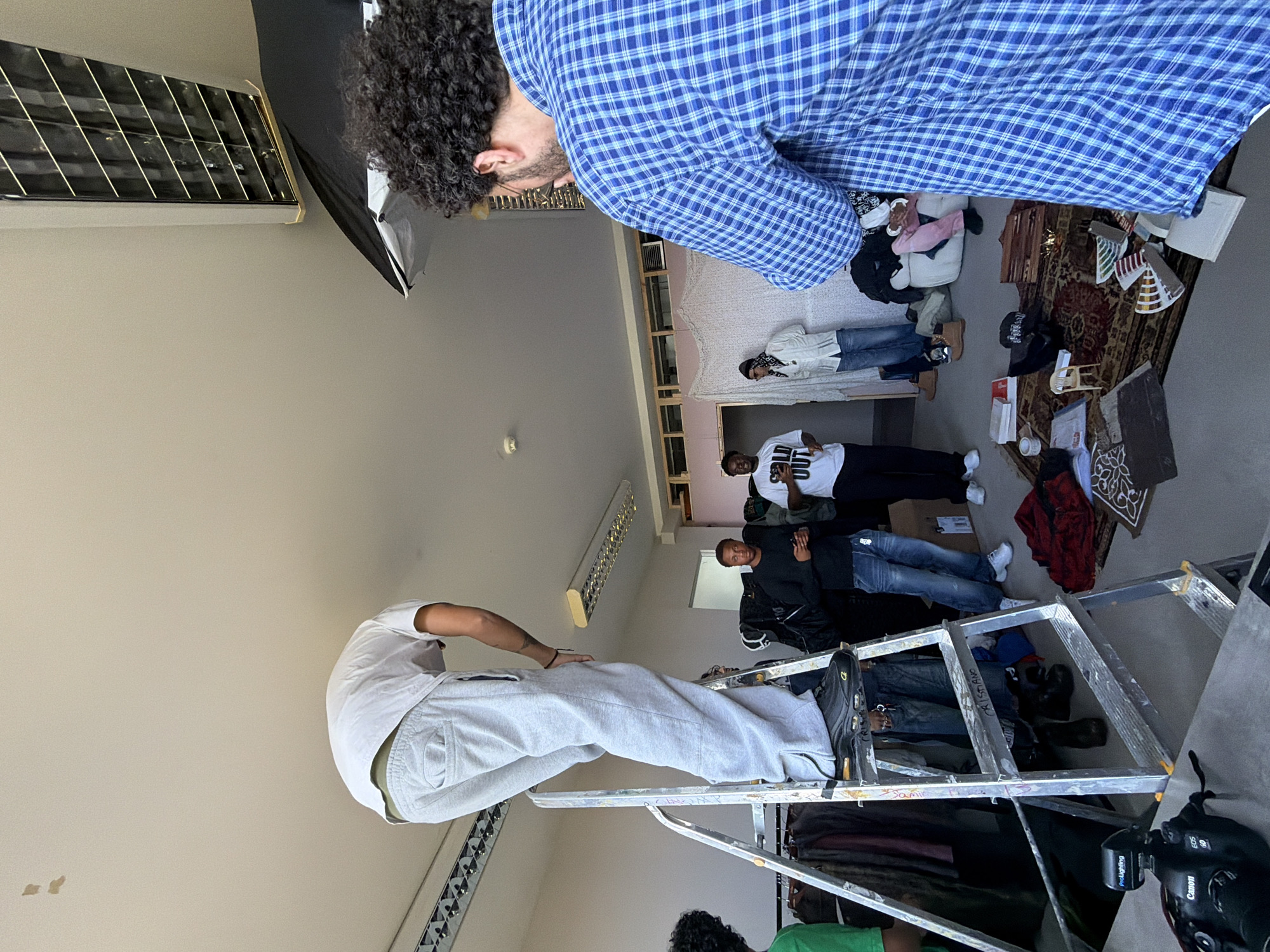 This image captures an indoor scene, likely a photoshoot or media production in a studio or large room in London, United Kingdom. The space features plain, light-colored walls and a ceiling with a long, rectangular light fixture and what appears to be an air conditioning unit on the left.
In the foreground, a person with dark curly hair, wearing a blue plaid shirt, is seen from behind, bending over and looking down towards the activity. Centrally positioned, another individual in a white T-shirt and light grey sweatpants stands on an A-frame ladder. The ladder itself bears handwritten markings such as "SAMI" and "CALI STEFANO."
Several other people are scattered across the midground and background, observing or participating. One person wears a white T-shirt with "OUT" visible on it, while another is dressed in a white jacket and blue jeans. The floor in the background is covered with patterned rugs and various items including clothing, shoes, and boxes, hinting at styling choices or props. A clothes rack is also visible on the far left.
A black Canon EOS R camera equipped with a "ProLighting" flash attachment is visible on the floor near the ladder, confirming the presence of a professional setup. The scene is well-lit, suggesting a combination of natural light and studio lighting from a softbox visible on the left. The overall mood appears focused and busy, with participants engaged in the creative process.