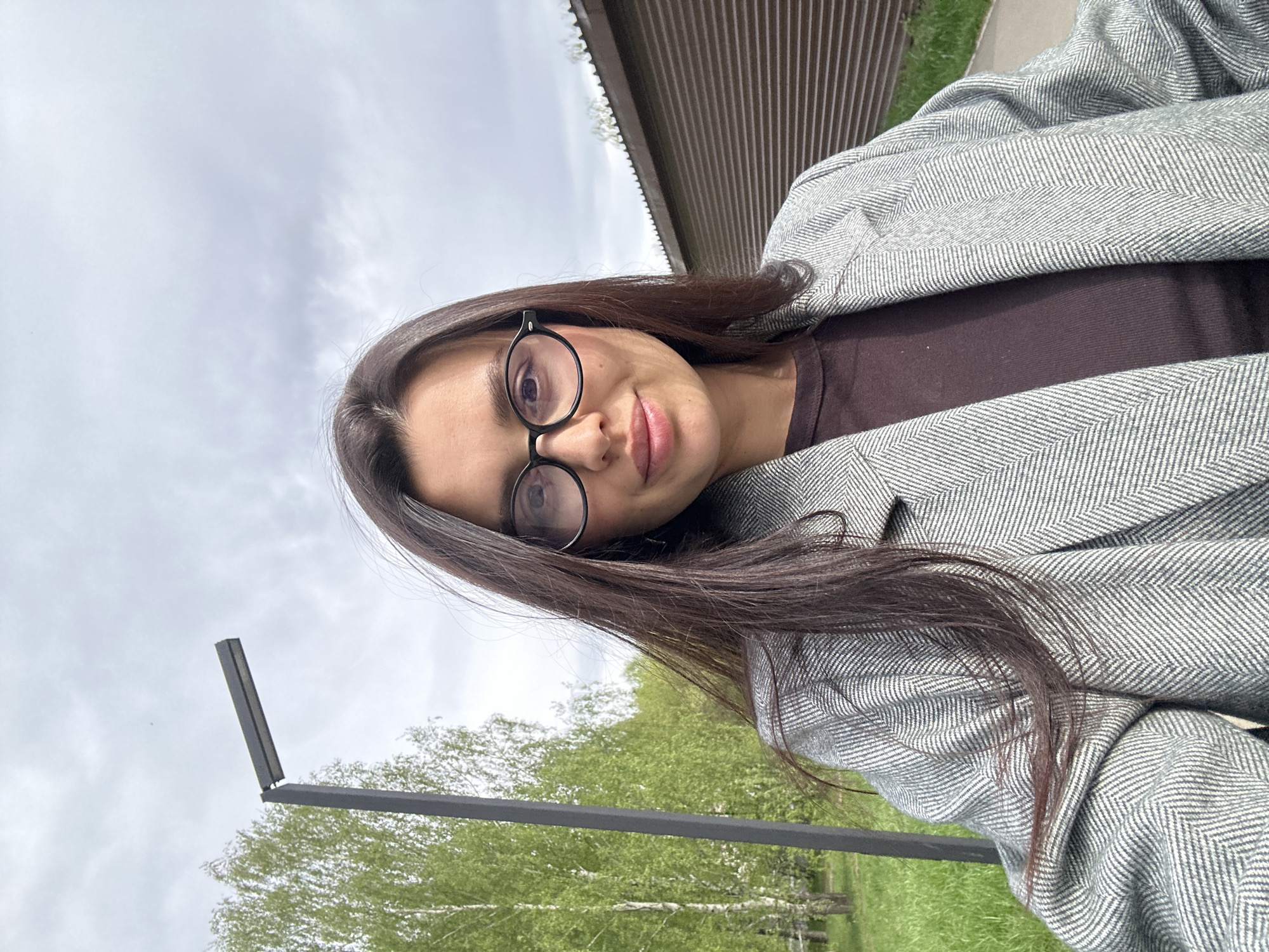 The image is a selfie of a woman with long brown hair and glasses. She is wearing a gray herringbone jacket over a brown top. The background includes a cloudy sky, trees with green leaves, a gray metal structure (likely a lamppost), and a building with dark brown siding. Based on the information about the location being Chaikovsky, Russia, it can be inferred that this is a park or outdoor area within the city.