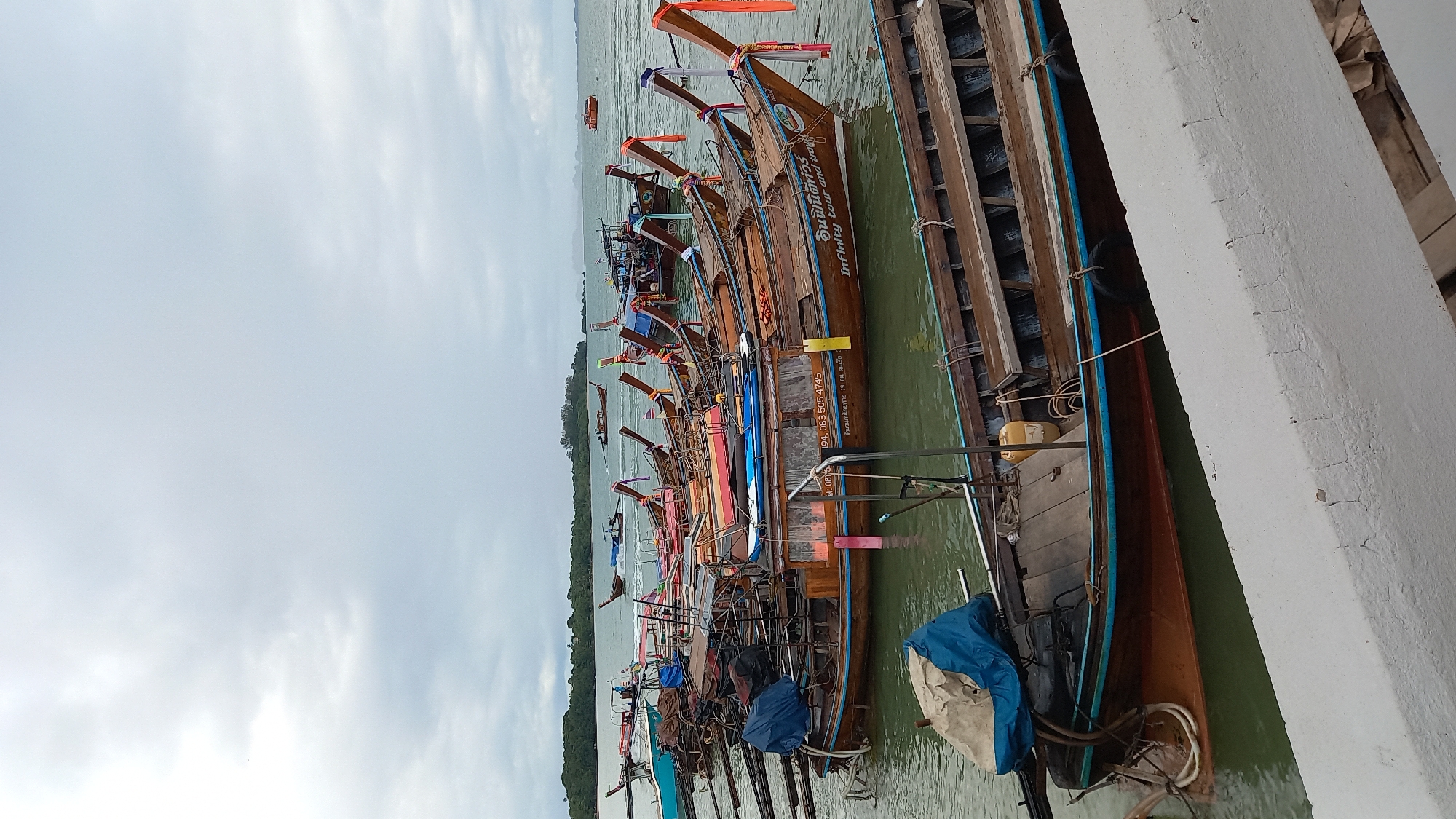 The image depicts a row of longtail boats docked in the city of Ao Nam Mao, Thailand. The boats are of varying designs, with some featuring wooden hulls and others painted in bright colors. Many of the boats have decorative prows and are adorned with flags or ribbons. The water is calm, and the sky is overcast. A concrete structure is visible on the right.