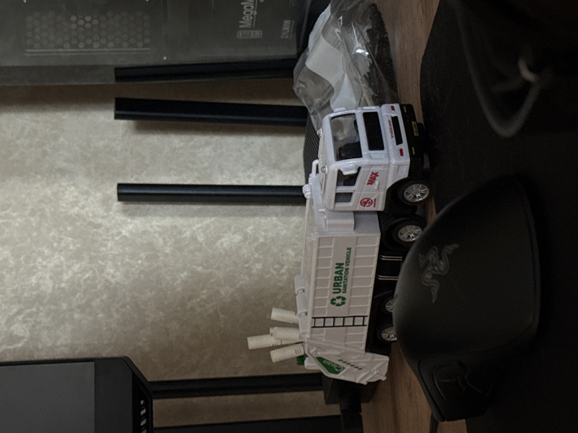 A sideways close-up shot shows a white toy garbage truck with "URBAN SANITATION VEHICLE" written on its side. It sits on a wooden surface next to a black computer mouse. Adjacent to the truck is some computer hardware. The photo does not suggest any identifiable location in Borzya, Russia.