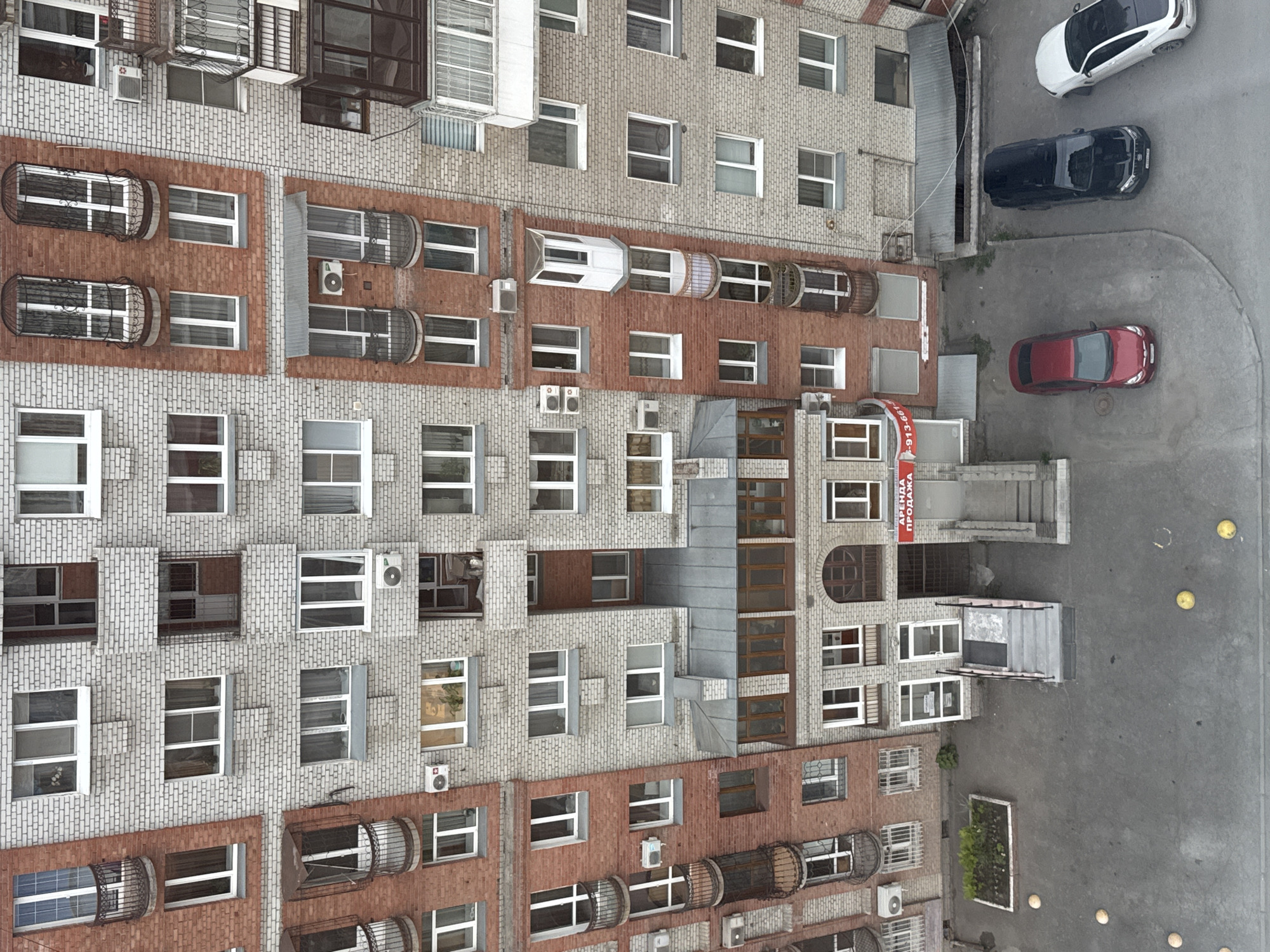The image shows an aerial view of a multi-story building in an unknown city. The building facade is a mix of gray and red brick, with numerous windows, some with decorative metal cages. Several air conditioning units are attached to the exterior walls. Next to the building, there's a small parking area with three cars: a white one, a black one, and a red one. Adjacent to the parking area is a section of pavement with a few yellow bollards, and a small planter with greenery. The scene suggests an urban environment, potentially a residential or mixed-use area.