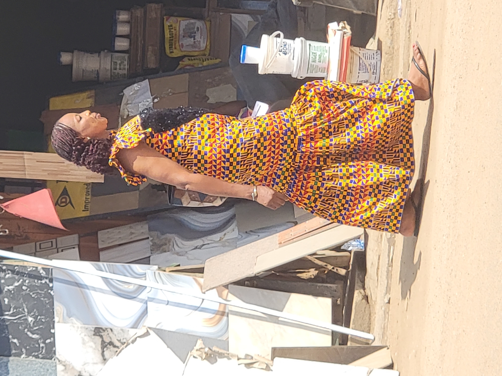 A middle-aged woman wearing a traditional African dress is standing on a sidewalk in Maiburiji, Nigeria. The dress is predominantly yellow and orange with a geometric pattern of blue and purple squares. She is also wearing flip-flops and a bracelet on her left wrist. Her hair is styled in small, dark braids. She appears to be looking towards the left, possibly observing something off-camera.
Behind her is a shop with stacks of tiles and construction materials. There are various boxes, buckets, and bags of cement or adhesive visible on shelves and on the ground. The shop front is somewhat cluttered, suggesting a busy retail environment.
The sidewalk is a light brown color, indicating it may be made of concrete or a similar material. There is also some debris or loose items scattered along the edge of the sidewalk. The lighting in the scene suggests it is daytime with bright sunlight.