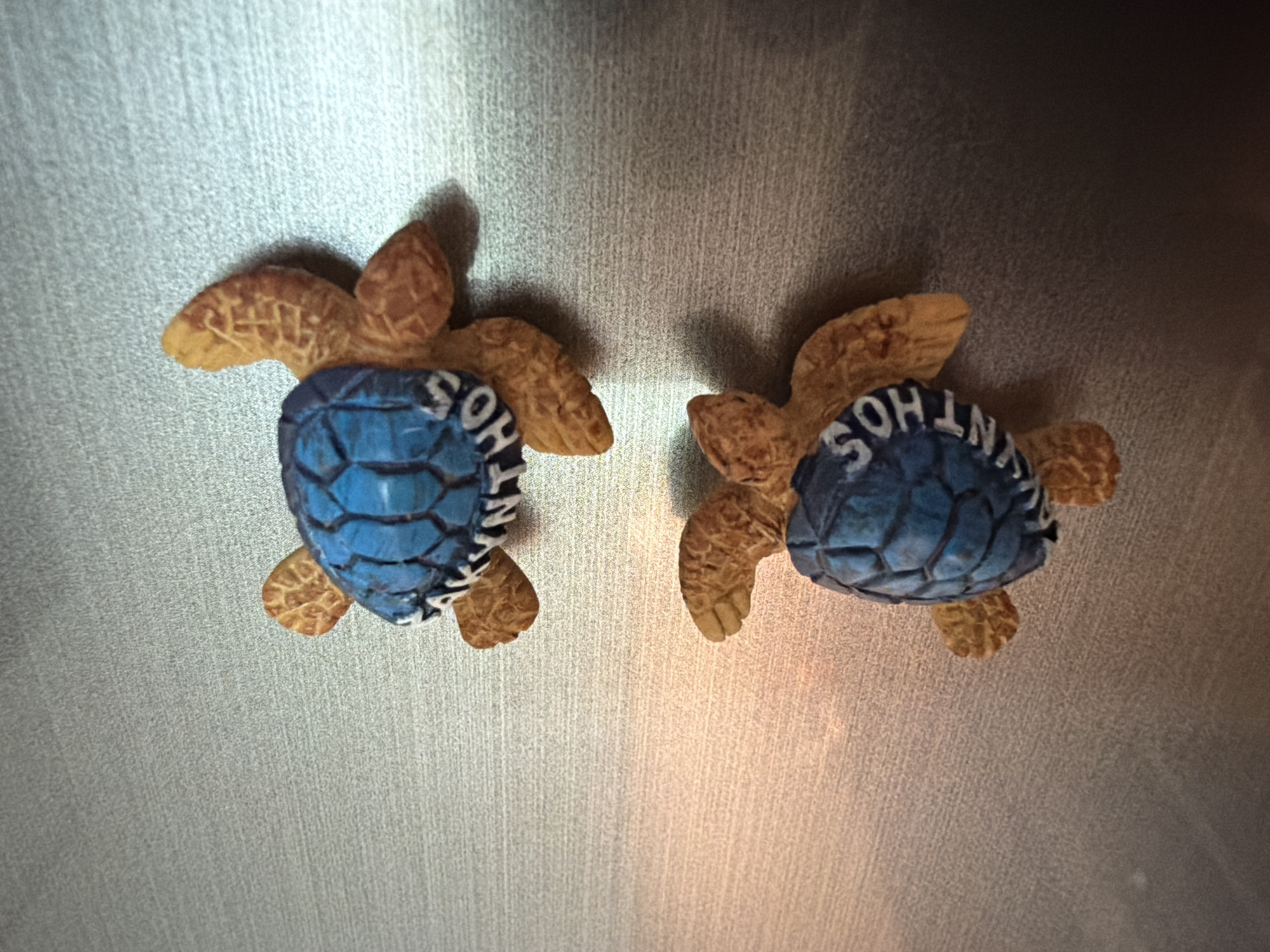 The image captures two decorative turtle-shaped magnets affixed to a brushed metallic surface, likely a refrigerator door, in Lusail, Qatar. Both turtles are identical, featuring a light brown, textured body and flippers, contrasted with a vibrant blue shell. The shells are segmented in a hexagonal pattern, and each bears the white capital letters "SOHNTOS" prominently inscribed on its surface.
The metallic background exhibits fine vertical brushing, creating a subtle texture. Shadows are cast by the turtles, predominantly to their left, suggesting a light source from the upper right. The lighting highlights the metallic sheen of the surface, which ranges in tone from bright silver on the left to a slightly warmer, darker gray on the right. The scene depicts a static display of objects, with no human activity or specific time of day evident beyond indoor lighting conditions.