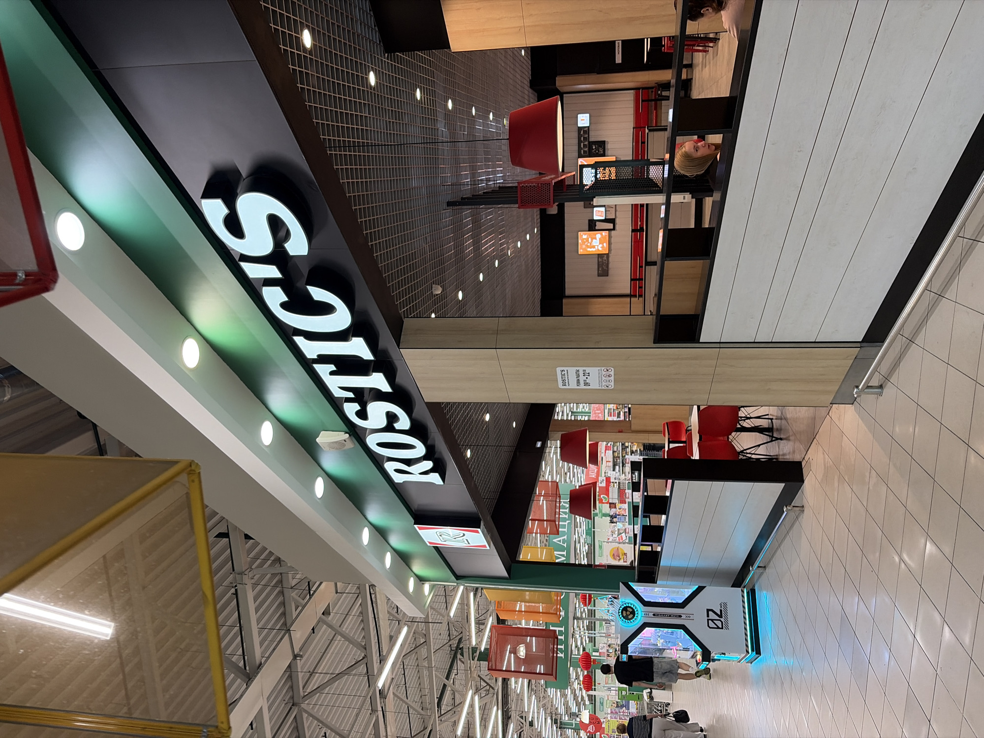 The image shows the interior of a Rostic's restaurant in Noginsk, Russia. The Rostic's sign is prominently displayed above the entrance. Inside, there are tables and chairs for customers, as well as food displays and a counter. The lighting is bright and modern, with a mix of overhead lights and pendant lamps. The floor is tiled, and the walls are decorated with colorful graphics. There are a few customers visible in the background, as well as a staff member behind the counter. The overall impression is that of a clean, modern, and inviting fast-food restaurant.