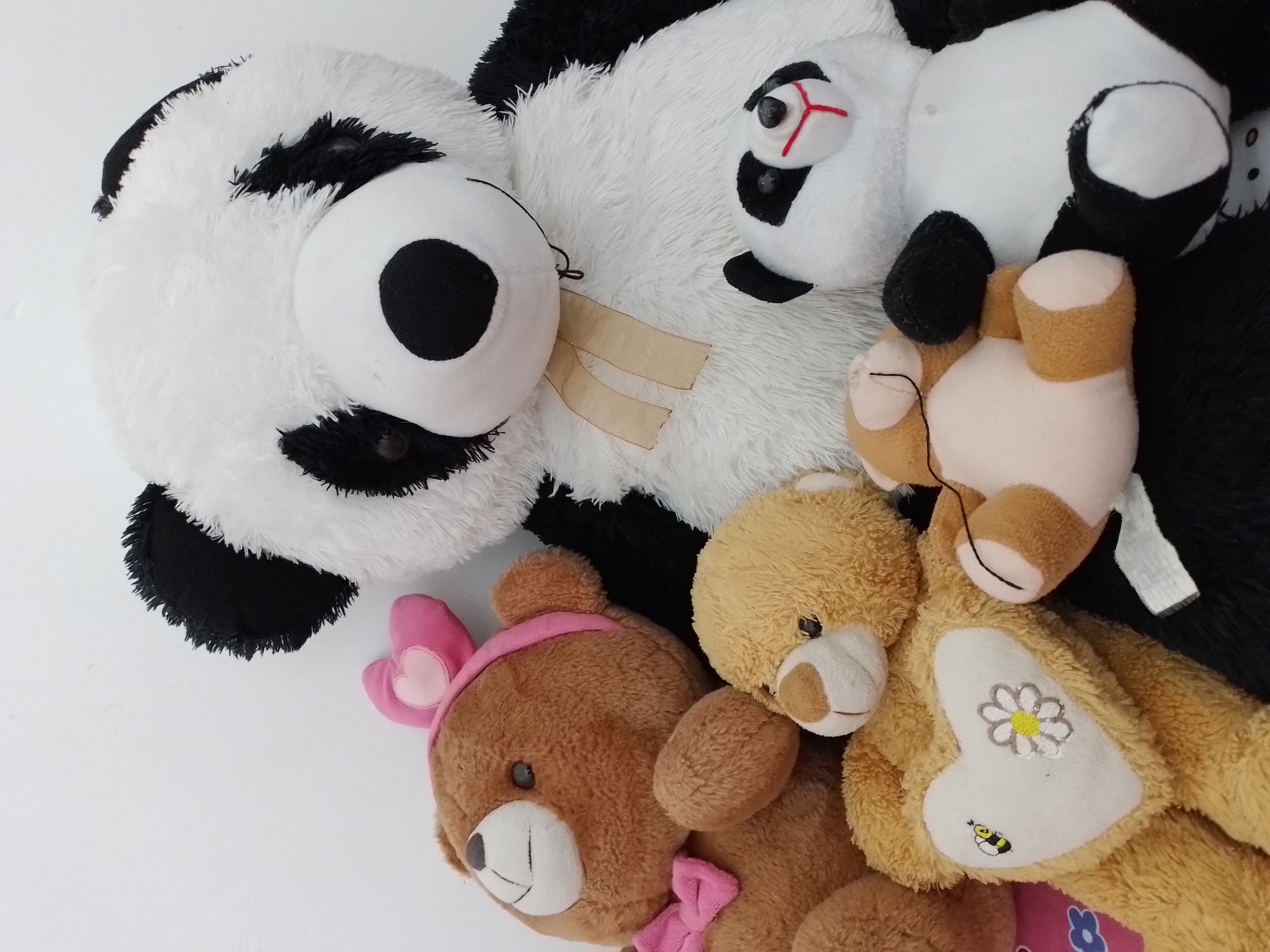 A group of plush toys is arranged against a white background. The toys include a large panda, a brown teddy bear with a pink bow and headband, a smaller yellow teddy bear with a heart on its chest adorned with a bee and flower, a tiny panda, and a small brown and white teddy bear. The panda toys are positioned in the center and right of the frame, while the teddy bears are on the left and bottom. The overall scene is static and appears to be a collection of stuffed animals.
