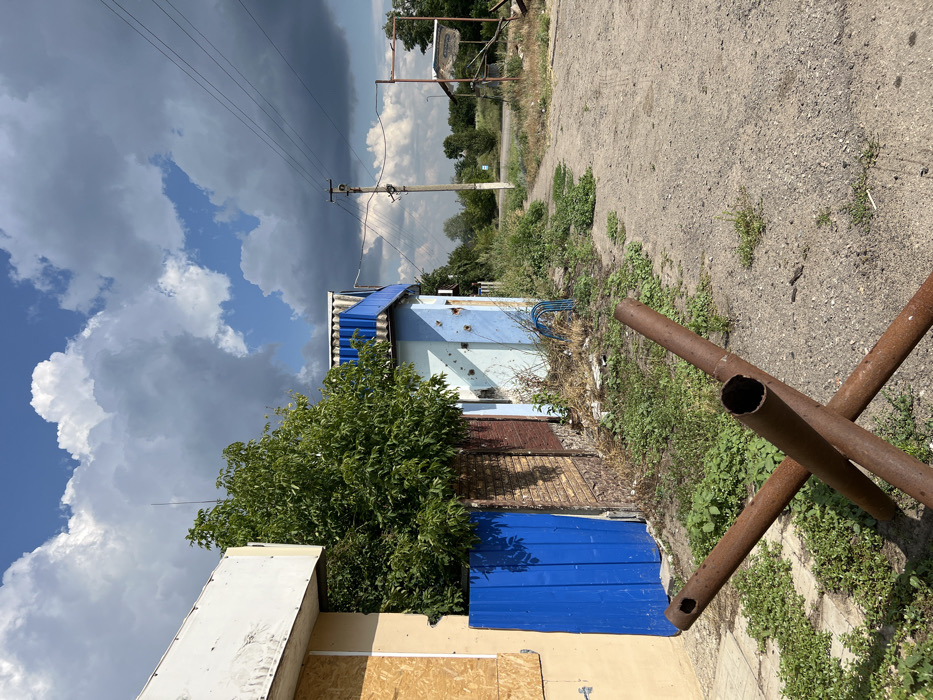 This is an outdoor daytime photograph depicting a damaged urban environment in Shestakove, Ukraine. In the immediate foreground, a rusty, brown anti-tank hedgehog, constructed from three intersecting hollow metal pipes, rests on an unpaved dirt and gravel path heavily interspersed with green vegetation and weeds. To the left, a building with a light-colored facade shows evidence of damage, with windows boarded up by light-brown plywood sheets. Adjacent to this structure is a solid blue metal gate or fence section, followed by a low brick wall. Behind these barriers, a dense green tree with numerous leaves is visible.
To the right of the boarded-up building, a single-story structure painted light blue features a prominent blue-trimmed corrugated roof and exhibits multiple circular impact holes on its exterior wall. The ground around this building is also overgrown with weeds and some debris. Further along the unpaved path, which curves slightly to the right, a utility pole with multiple overhead power lines stands upright. In the mid-ground to background, another dilapidated structure or metal frame is visible to the right of the path, with additional green vegetation and trees defining the distant horizon. The sky overhead is partially cloudy, displaying large, white cumulus clouds against patches of blue, with darker grey cloud formations present, particularly towards the right side of the frame. No individuals are visible.