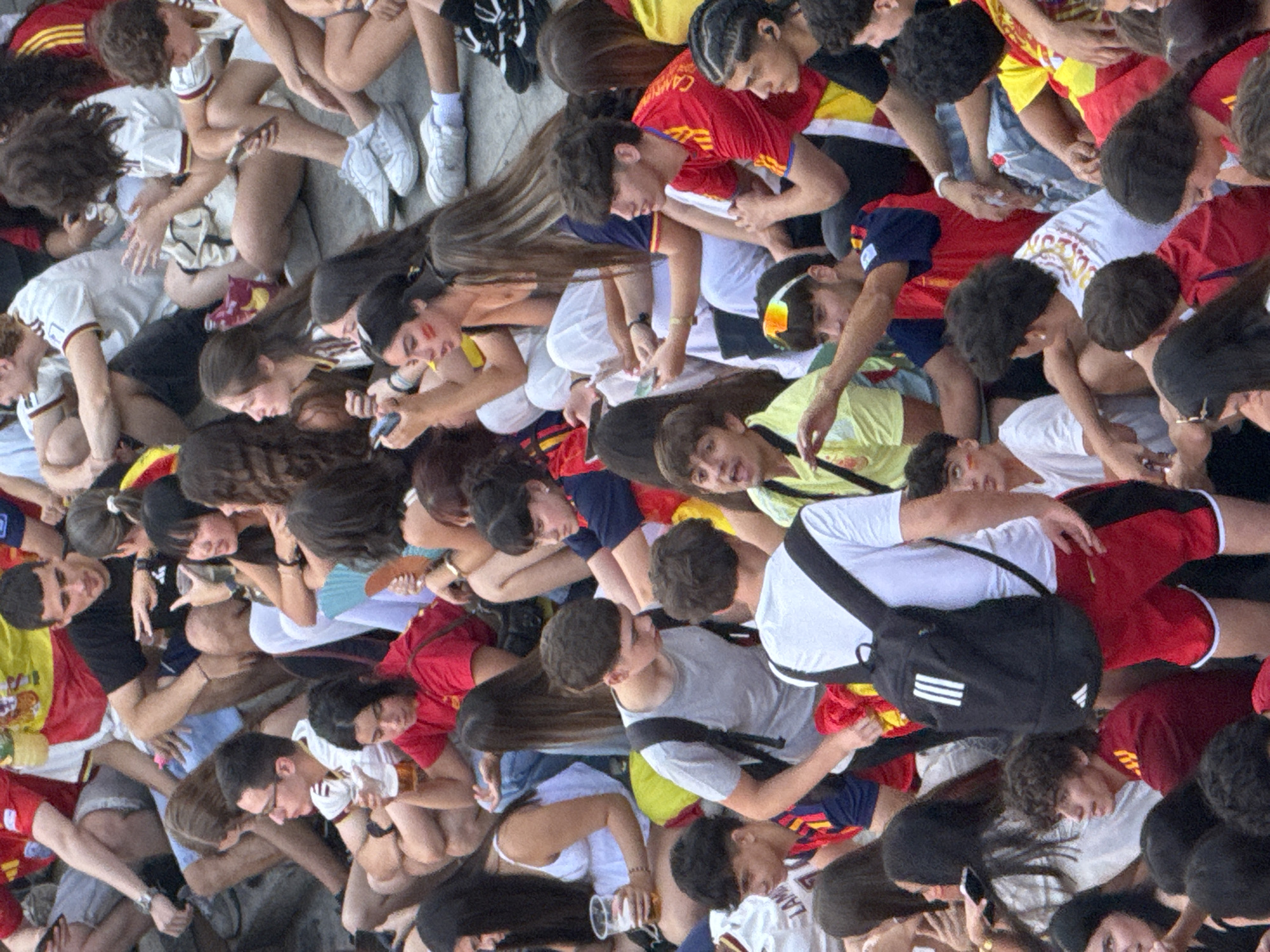 The image depicts a crowd of people, likely gathered for an event, on a paved surface, possibly a stadium or public square, in Las Rozas de Madrid, Spain. The individuals, predominantly young adults and teenagers, are seated or standing, many wearing sportswear in red, yellow, and blue, suggesting a sporting event or national celebration. Several people are holding drinks and looking in various directions, indicating a casual and engaged atmosphere. Some are interacting with their phones, while others are observing the surroundings. The lighting suggests it is daytime, possibly late afternoon or early evening, with natural light illuminating the scene. The overall impression is one of a lively and communal gathering.