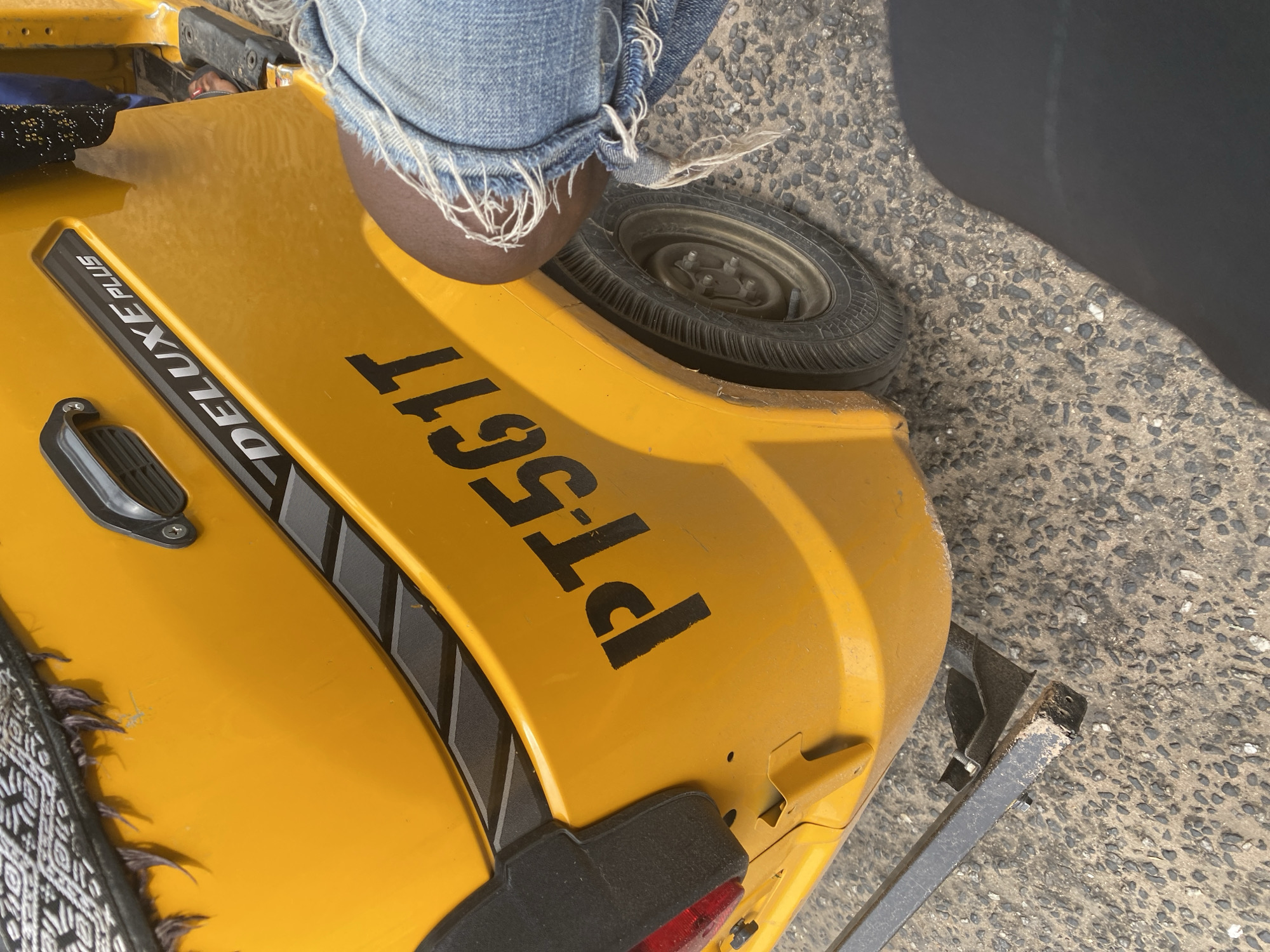A person wearing distressed denim shorts sits on a bright yellow three-wheeled vehicle. The vehicle has "DELUXE PLUS" printed on its side in a silver and black font. The license plate number "PT5611" is clearly visible in large black lettering on the vehicle's rear. The scene appears to be outdoors, with a rough, gravelly surface visible beneath and beside the vehicle. The lighting suggests it is daytime, possibly with strong sunlight casting a shadow on the ground. The person's legs are partially visible, with the frayed edges of their shorts and a glimpse of their knee. The overall impression is a casual, everyday scene in a location like Jalingo, Nigeria.