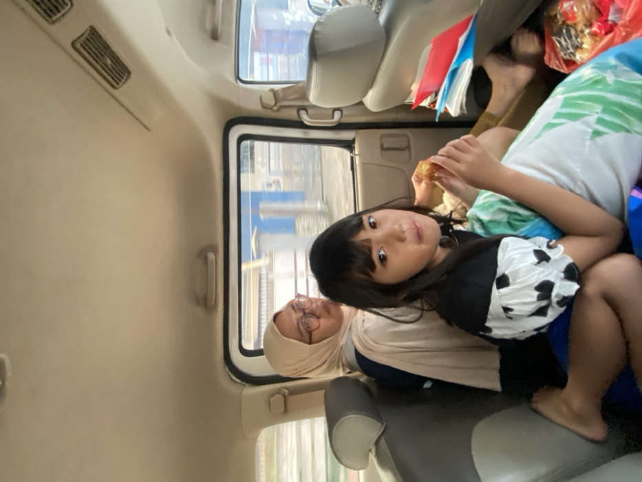 Inside a vehicle, a young girl with bangs sits beside a woman wearing a hijab. The girl looks directly at the camera. She wears a black top with puffy, spotted sleeves. Her mother is looking out the window. Another person's bare feet are visible toward the top right of the frame, suggesting a crowded space. A window shows outside scenery passing by. There appear to be food items and travel supplies in the back.