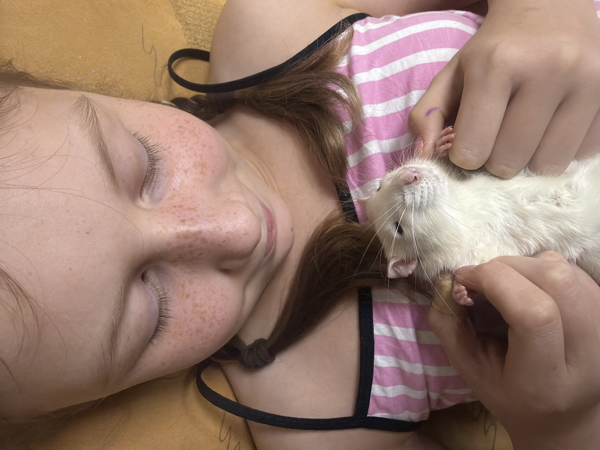 A young girl with light skin and freckles is lying down, eyes closed. She is wearing a pink and white striped tank top with black straps. Her light brown hair is pulled back in two small ponytails. She is holding a white rat. The background appears to be a yellow or gold blanket or sheet. The location is not evident based on the image.