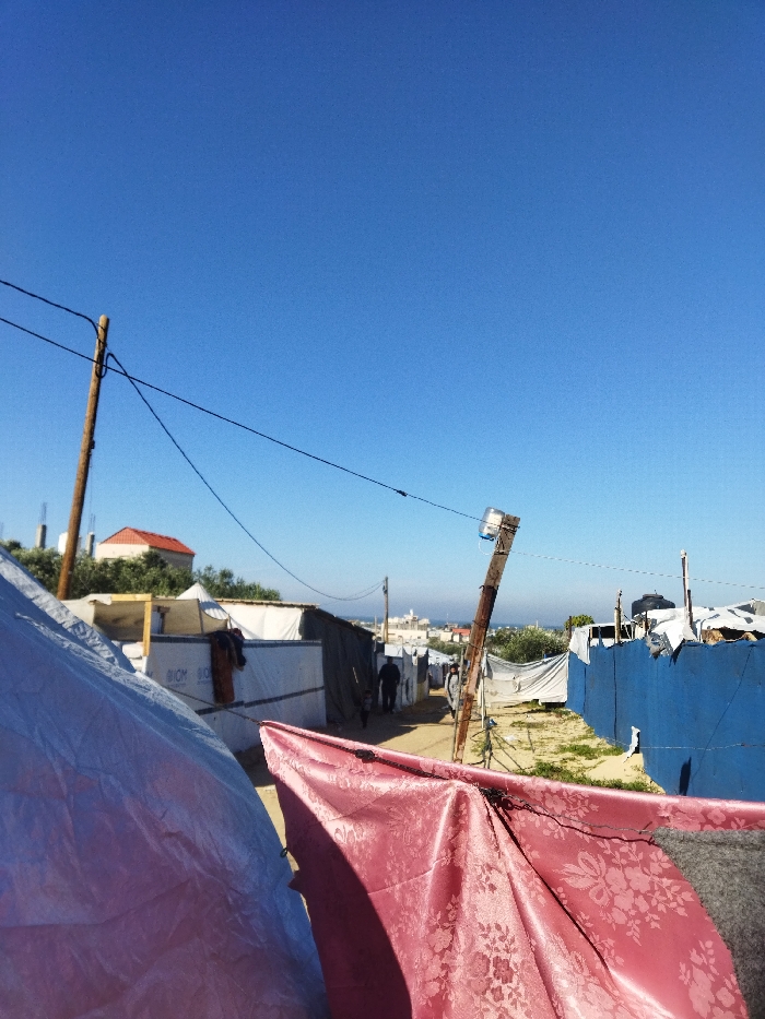 The image depicts an outdoor scene within a makeshift settlement, likely located in Khan Yunis, Palestinian Territory, under a clear blue sky. A narrow dirt path bisects the view, receding into the distance. Along both sides of the path are temporary shelters constructed from diverse materials including white panels (some branded "BIOM"), tarpaulins, dark fabrics, corrugated sheets, and wooden supports. Clothes are seen hanging outside one shelter on the left. Two wooden utility poles stand along the path; one on the left with overhead wires, and another leaning significantly to the right, also carrying wires, with a clear plastic bottle affixed near its top. Two individuals, an adult-sized figure and a smaller figure, walk away from the viewer down the path in the mid-ground. In the left background, a building with a reddish-orange tiled roof is visible on a rise, amidst green foliage. The distant horizon features a hazy outline of structures and a bright, indistinct line, possibly indicating a coastal area or distant cityscape.