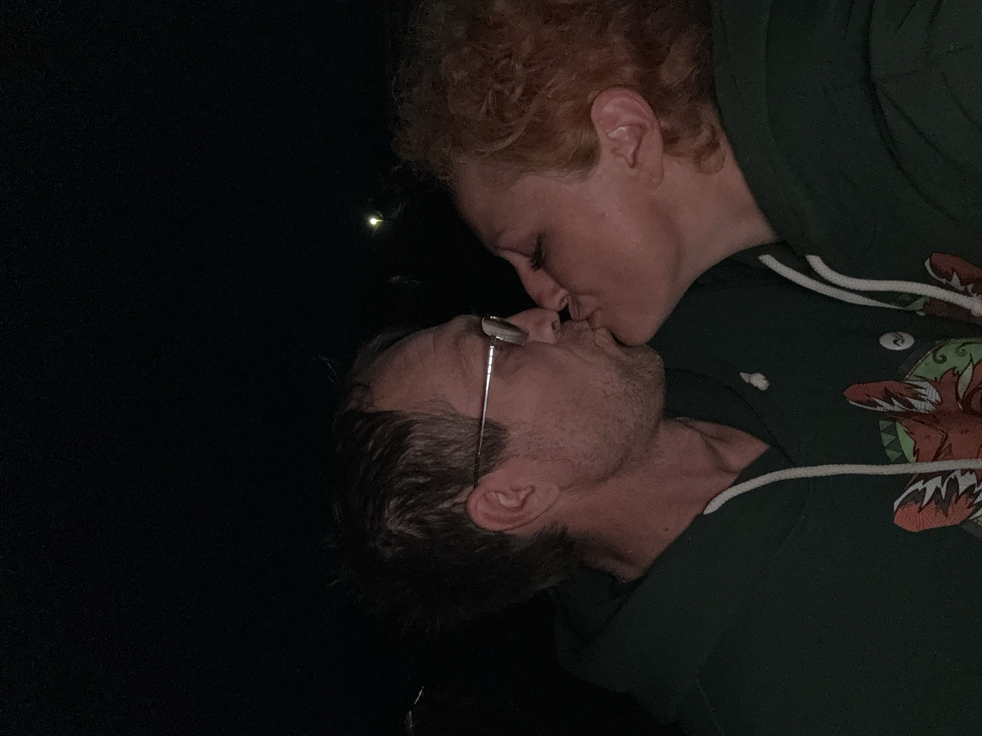 The image depicts two individuals, a man and a woman, engaged in a kiss in a dimly lit setting, likely at night, in Большая Кивара, Russia. The man, on the left, has short, textured hair, facial stubble, and wears eyeglasses. He is dressed in a dark green hooded sweatshirt with white drawstrings and a visible graphic on the chest featuring a stylized animal face, primarily in orange and white tones. The woman, on the right, has curly, reddish-brown hair and is also wearing a dark green hooded sweatshirt. Their faces are in profile, with their lips pressed together. The background is largely dark, with a singular, bright light source visible in the upper-right portion of the frame.