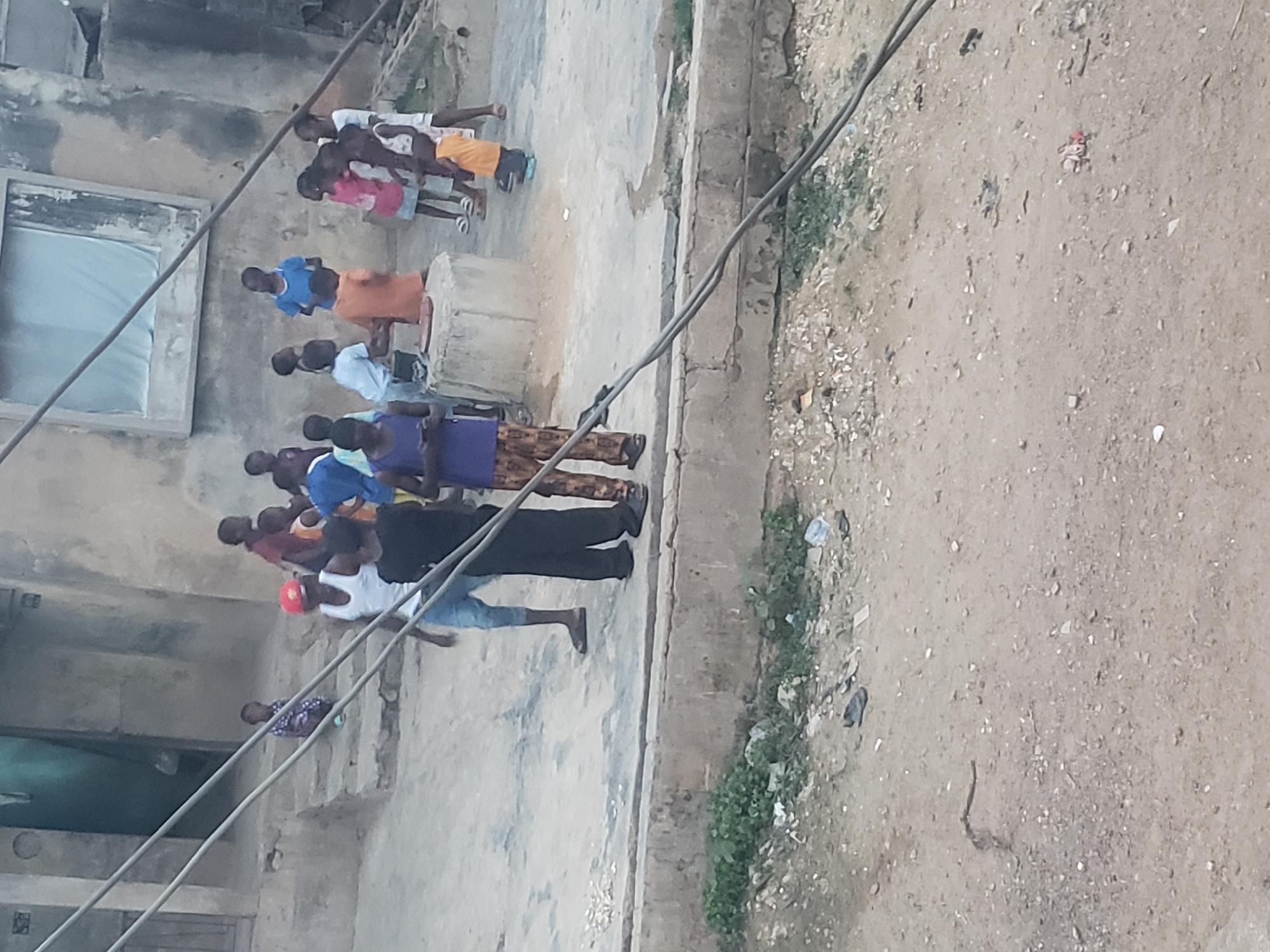 A group of people stand on a street in Alalubosa, Nigeria. The street is bordered by buildings and a dirt area with vegetation. The people appear to be observing something, possibly performing an action. Power lines are visible in the foreground. Some litter is scattered on the ground.
