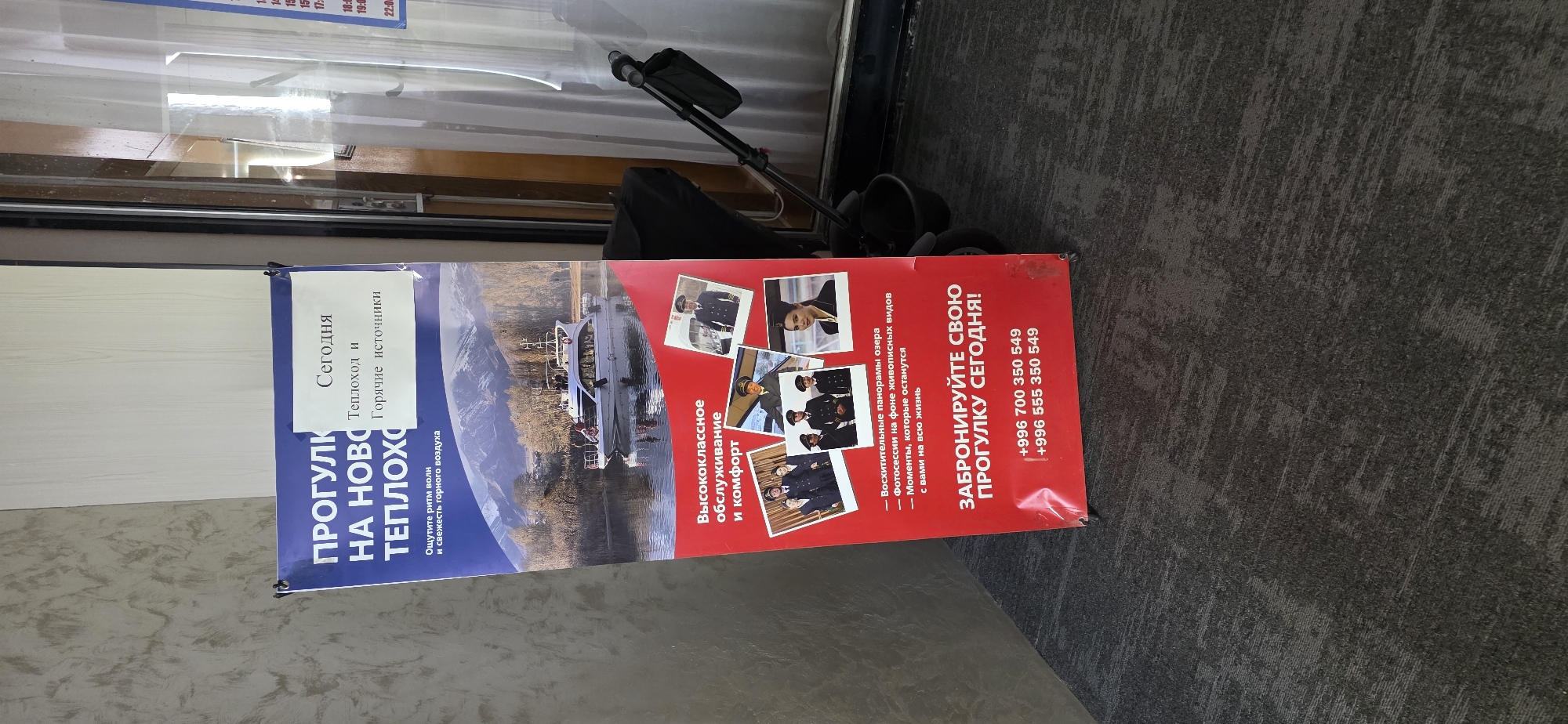 The image shows a vertical banner with text and images, likely an advertisement for a boat tour. The banner is positioned outdoors, possibly near a waterfront, with a grey, textured carpet visible in the foreground.
The banner's primary subject is a boat tour, indicated by the large text "ПРОГУЛК НА НОВО ТЕПЛОХО" which translates to "WALK ON NEW STEAMSHIP" or "CRUISE ON NEW STEAMSHIP." The text further elaborates on the experience, mentioning "Теплоход и Горячие источники" (Steamship and Hot Springs), and "Ощутите ритм волн и свежесть горного воздуха" (Feel the rhythm of the waves and the freshness of the mountain air).
The banner features several images: a large photograph of a boat on a body of water, likely a lake, with mountains in the background. Smaller inset photos showcase people in uniform, possibly crew members or participants, and a woman. Text on the banner highlights "Высококлассное обслуживание и комфорт" (High-class service and comfort) and promises "Восхитительные панорамы озера - Фотосессии на фоне живописных видов - Моменты, которые останутся с вами на всю жизнь" (Magnificent lake panoramas - Photoshoots against the backdrop of picturesque views - Moments that will stay with you for a lifetime).
A call to action is prominently displayed: "ЗАБРОНИРУЙТЕ СВОЮ ПРОГУЛКУ СЕГОДНЯ!" (BOOK YOUR WALK TODAY!). Below this, two phone numbers are listed: "+996 700 350 549" and "+996 555 350 549", with the "+996" country code suggesting the location is Kyrgyzstan.
The overall impression is that of a promotional advertisement for a scenic and comfortable boat excursion, likely in a mountainous region with hot springs. The lighting suggests it could be daytime.