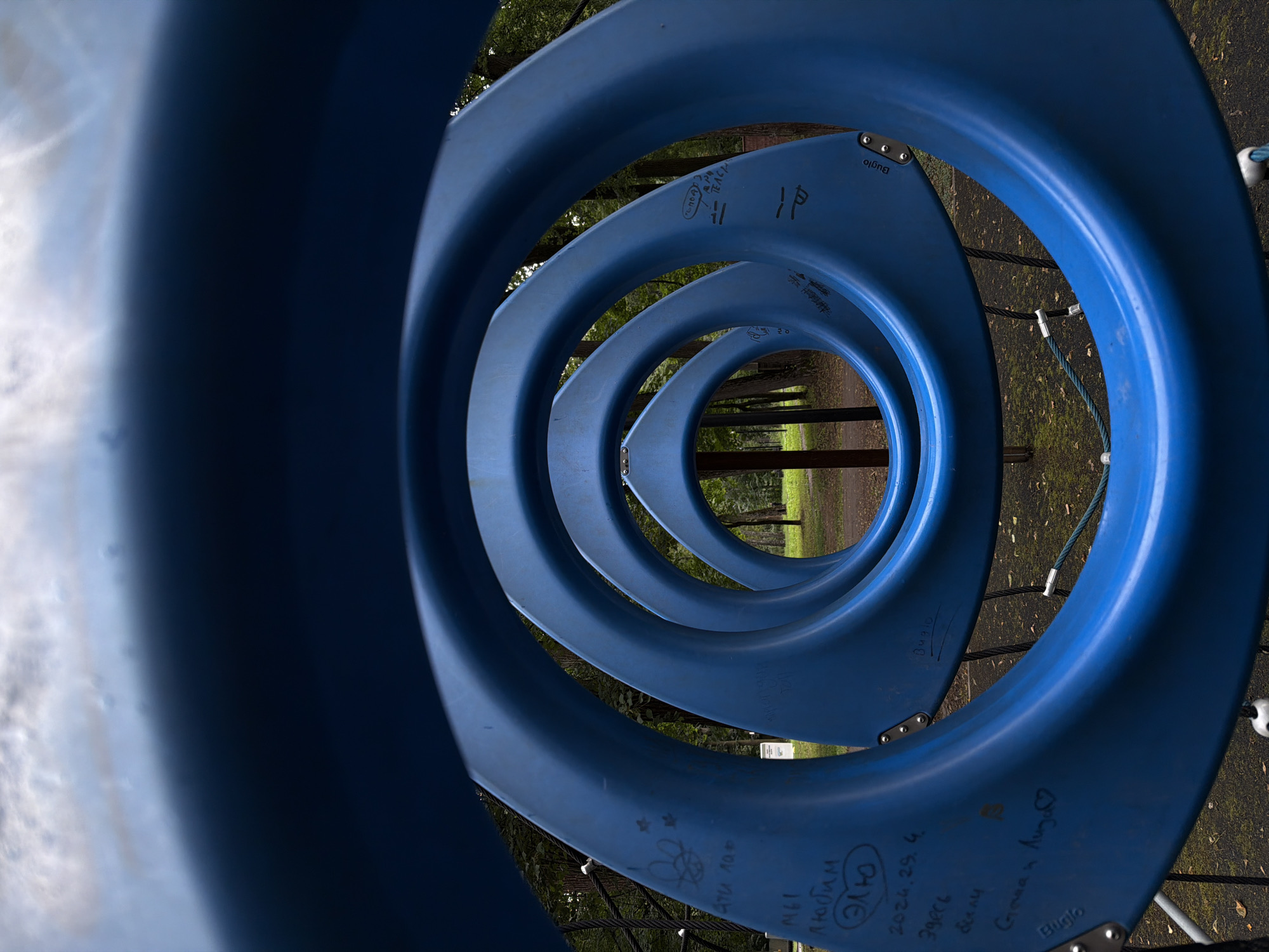 The image depicts a playground in Malakhovka, Russia. The foreground is dominated by large, curved, bright blue plastic structures, possibly part of a climbing or sliding apparatus. These structures form concentric rings, creating a tunnel-like effect that draws the viewer's eye inward towards the background.
The background reveals a park-like setting with trees, green grass, and fallen leaves on the ground. The sky is visible above, partially obscured by clouds. There are no people visible in the image.
Several instances of graffiti are present on the blue plastic surfaces. On the closest blue structure, in Russian script, some of the visible markings include "эти лап" (these paws), "МЫ любим" (WE love), "Элю" (Alyu), "2024.29.4." (a date), "Здесь были" (Here were), and "Слава и Лиза" (Slava and Liza) with a heart symbol. Another structure has markings that include "ПАР" and what appears to be "Buglo" with a heart. Further in, on another ring, there are markings that appear to be "ПОД" (under) and "ТЕАСН" (teacher), along with a stylized "11" and "IP". The word "Buglo" is also visible on another part of the structure.
The overall impression is a somewhat weathered playground, with evidence of past use and personal markings etched into its surfaces. The perspective is wide-angle, emphasizing the curves of the playground equipment and the depth of the scene.