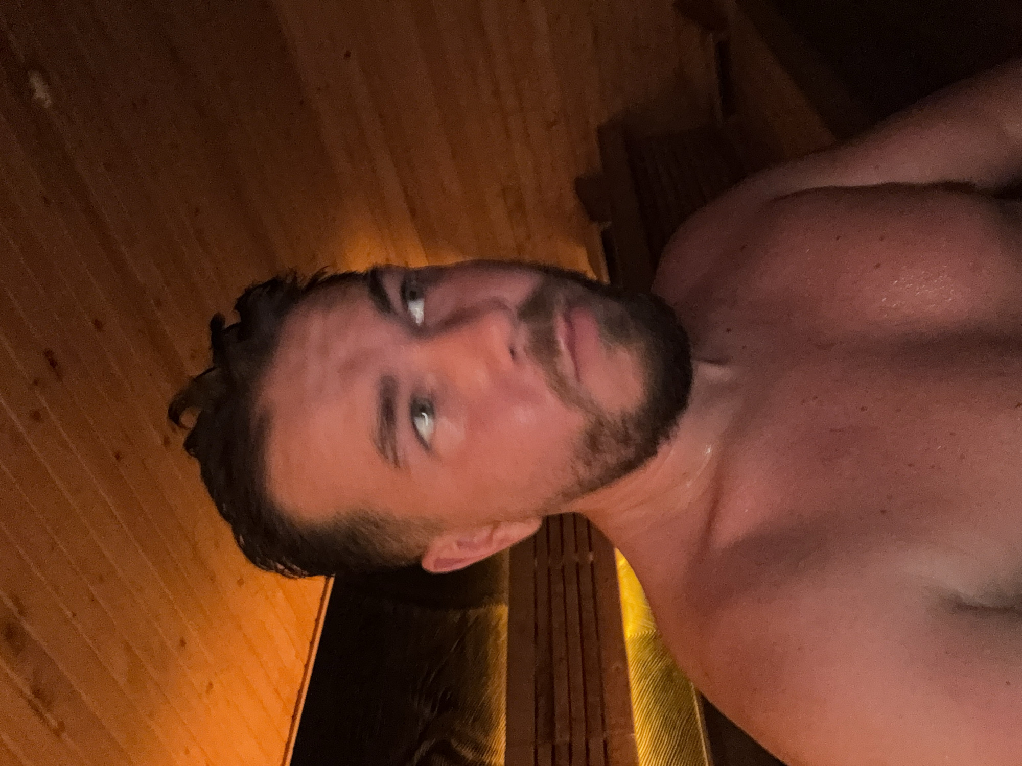 A close-up, angled shot of a man with short dark hair and a beard, appearing to be in a sauna. His skin is flushed, possibly from the heat. The wooden walls and benches of the sauna are visible in the background. The image contains no indicators regarding the location of Mengwi, Indonesia.