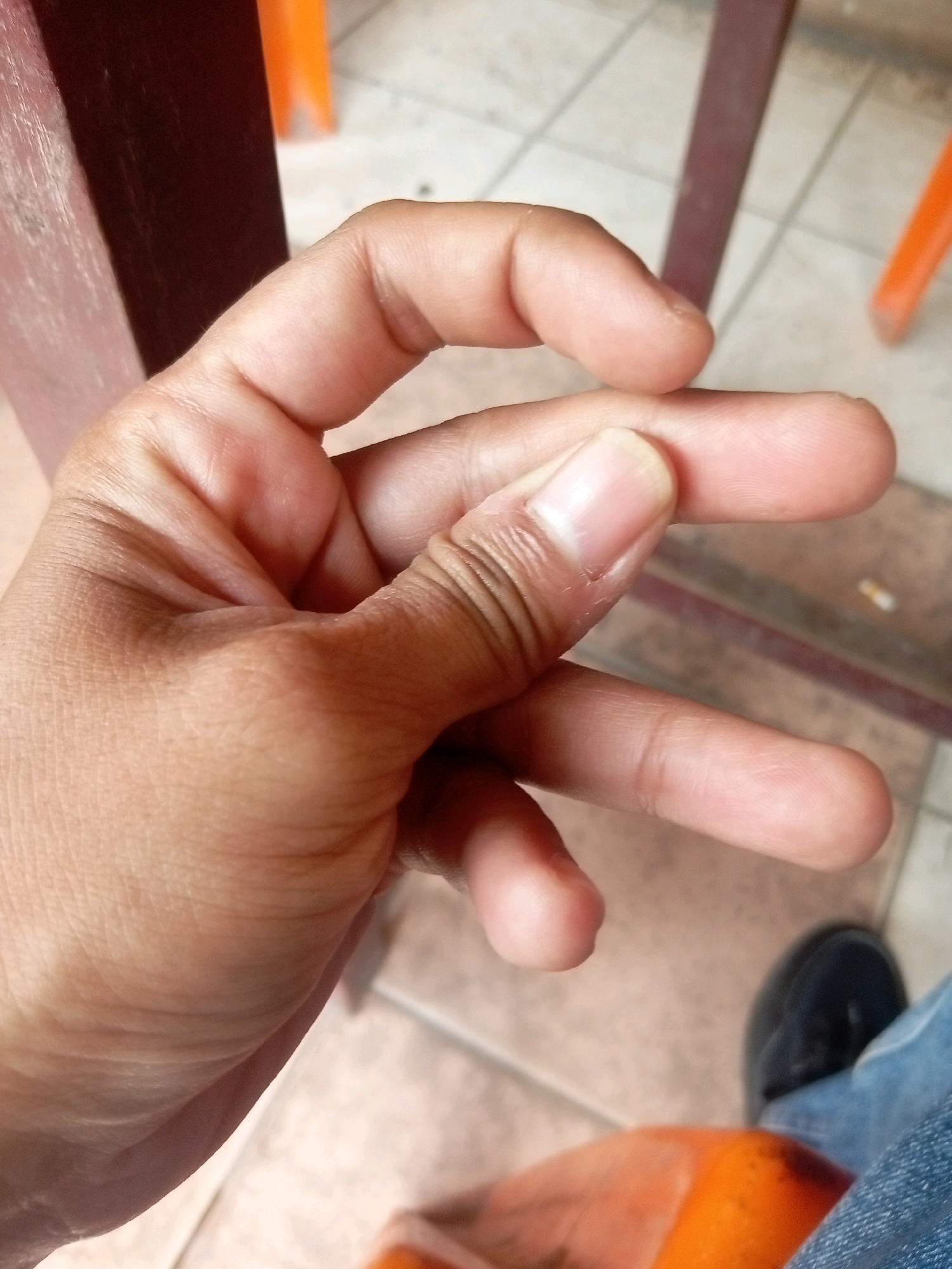 A close-up, high-angle shot focuses on a person's hand and fingers. The fingers are positioned in a way that suggests a gesture or a grip. The background is blurred, but visible elements include a tiled floor, a wooden chair or table leg, and what appears to be part of an orange plastic chair. The overall lighting is natural, with shadows indicating overhead illumination. The scene is an indoor setting, possibly a cafe or restaurant, based on the furniture and flooring.