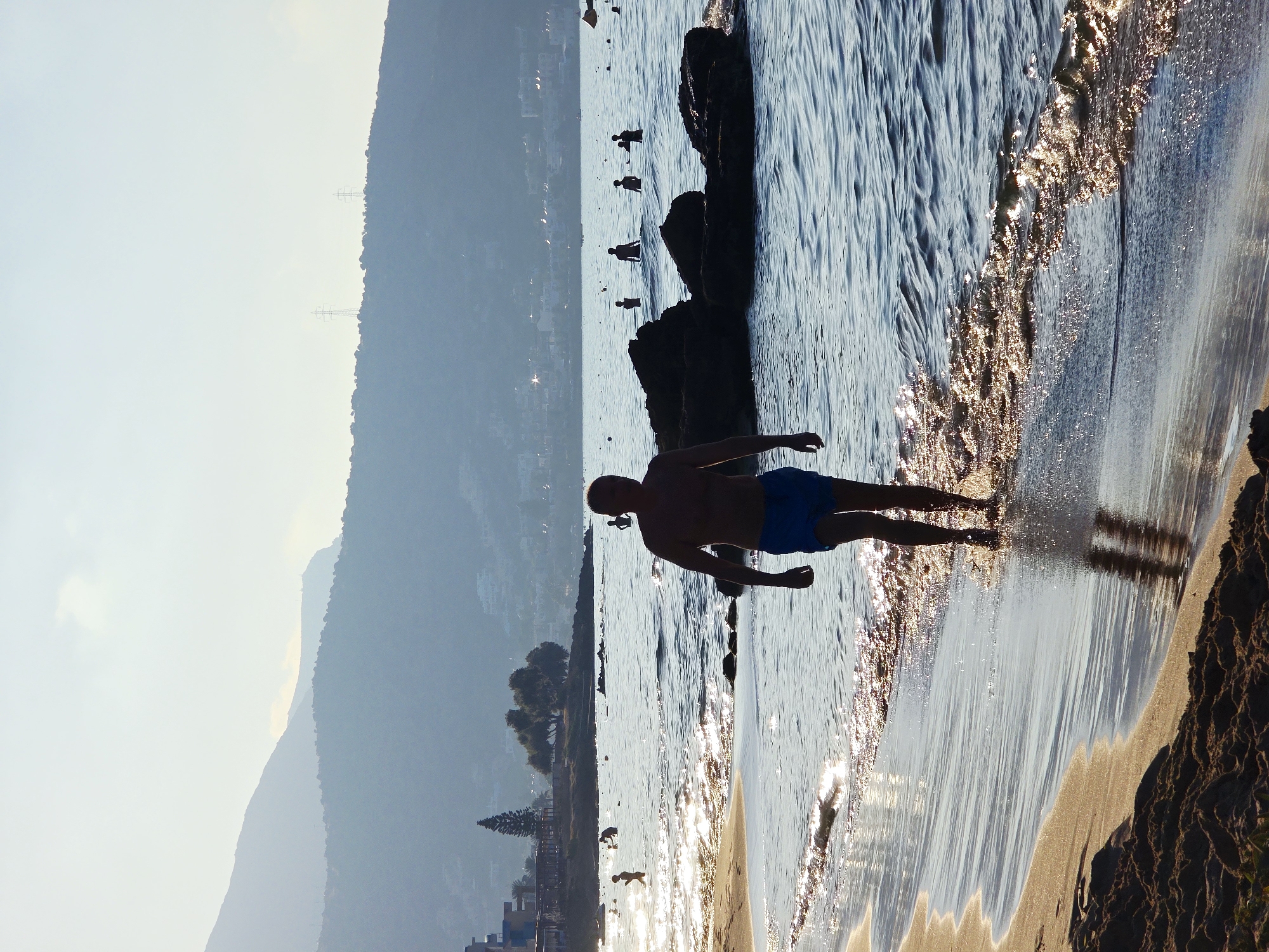 A silhouetted man in blue swim trunks walks along the wet sand of a beach in Malia, Greece. The sun is low in the sky, casting a warm light and creating shimmering reflections on the water. In the background, other people are visible wading and swimming in the sea. Rocky outcrops jut out from the water, and further in the distance, hills rise, with the outlines of buildings visible on their slopes. The sky is a pale, hazy blue. The scene conveys a peaceful, late afternoon or early morning atmosphere at the seaside.