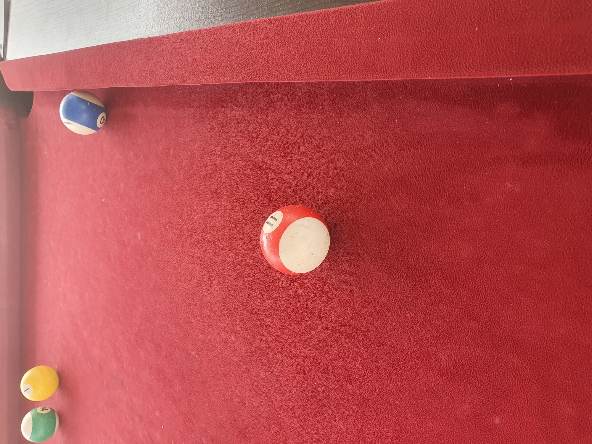 The image shows a close-up view of a red billiard table. There are four billiard balls visible on the table. In the upper left corner, there are two balls: one is green with a white stripe and the number 4, and the other is yellow with a white stripe and the number 1. To the right of these, further up the table, is a ball that is white with blue stripes and the number 0. In the lower center of the image, there is a red and white striped billiard ball with the number 11. The lighting suggests it is daytime, and the surface of the table appears slightly dusty. The image does not contain any elements that can be used to determine its location in Lokoja, Nigeria.
