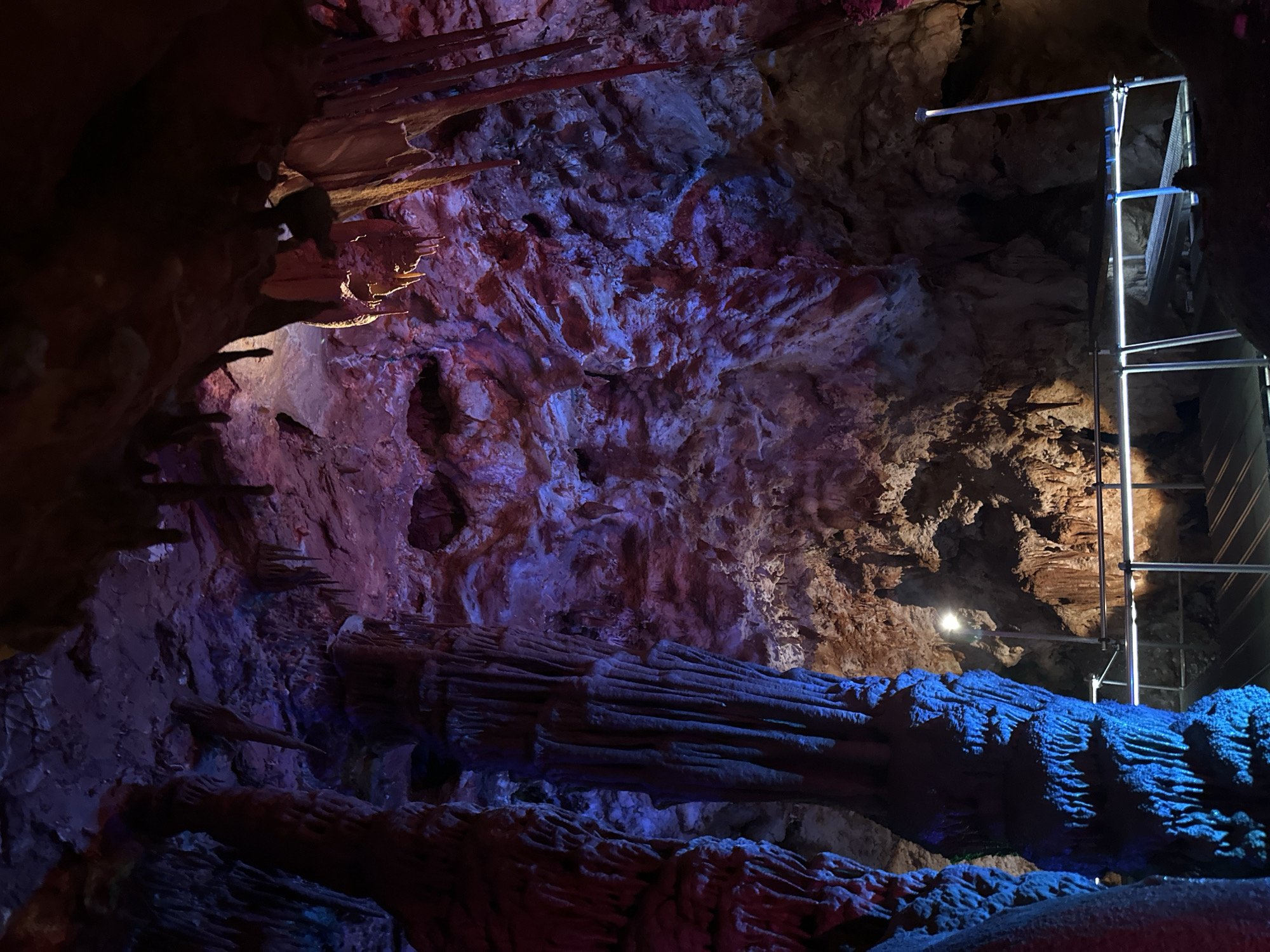 This image captures the interior of the Orlova Chuka cave, located in Belo pole, Bulgaria. The cave's interior is illuminated with colored lighting, predominantly in shades of blue, purple, and red. The geological formations of the cave, including stalactites and stalagmites, are prominently displayed. Scaffolding is visible on the right side of the image, indicating maintenance or exploration activities.