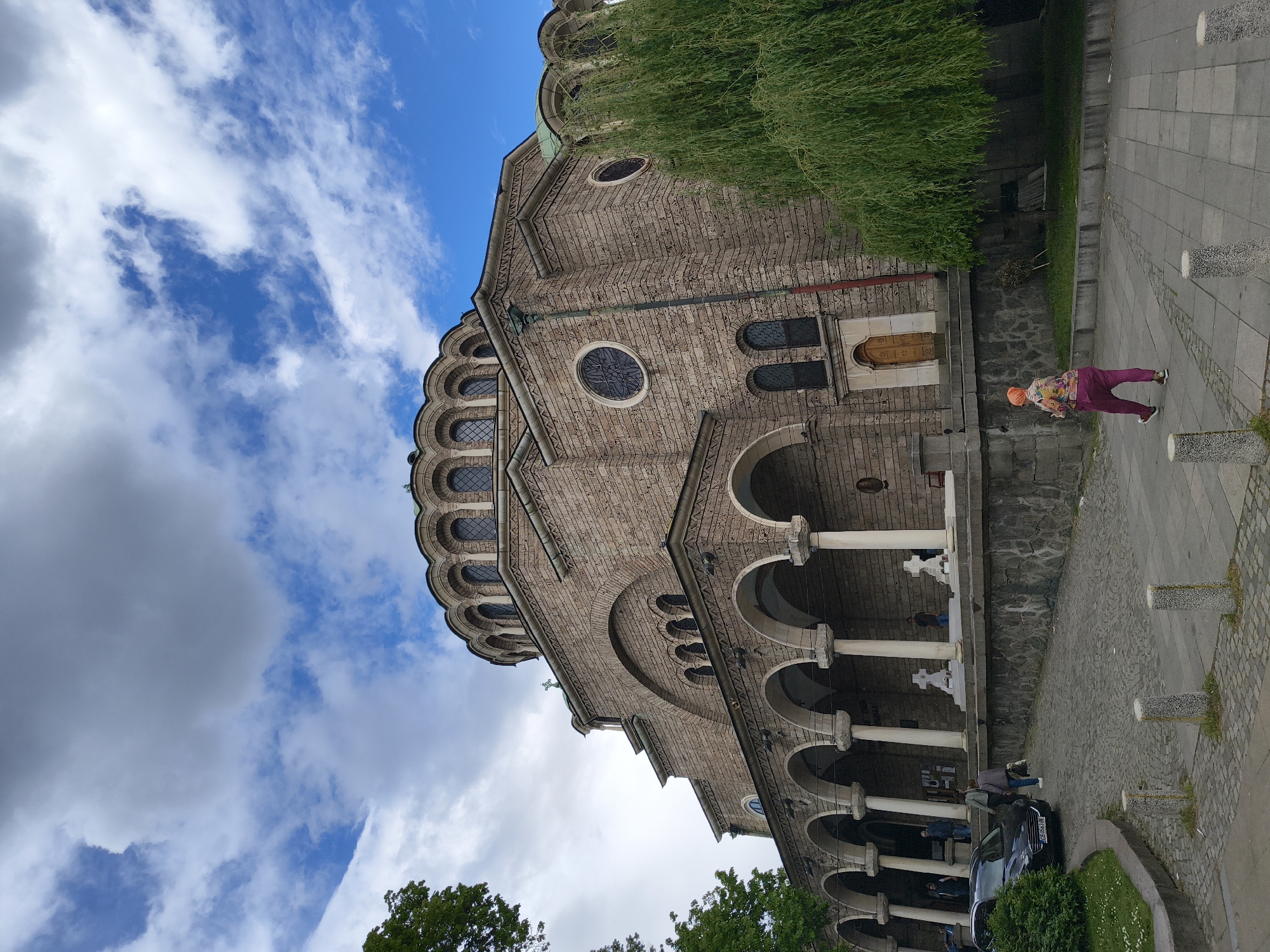 The image shows the Saint George Rotunda Church in Sofia, Bulgaria, from a low angle. The church is made of brick and stone, with a dome, arched windows, and a colonnaded entrance. The sky above is blue with white clouds. To the right of the church, there is a sidewalk made of cobblestones and gray concrete. An older woman dressed in purple pants, patterned top and an orange head covering is walking away from the camera. A car is parked in front of the church below the colonnade. A tree with green leaves is visible in the foreground on the bottom left.