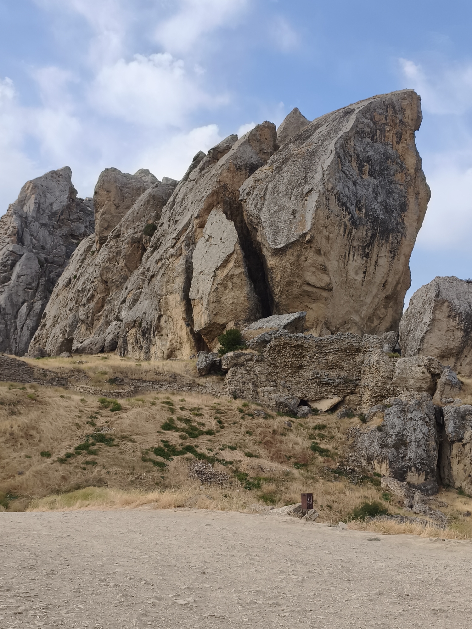 The image depicts a natural landscape dominated by large rock formations in Qalaşıxı, Azerbaijan. In the foreground, a dirt path with scattered pebbles leads into the scene. Behind the path, a dry, grassy hillside slopes upwards, dotted with sparse green vegetation and some scattered rocks. The middle ground features the base of imposing rock cliffs, with some evidence of ancient stonework, suggesting ruins or fortifications. These rocks are weathered and vary in color from light tan to dark gray. The upper portion of the image is dominated by the sheer, massive rock formations that rise dramatically against a bright blue sky with scattered white clouds. The overall scene conveys a sense of rugged, historical natural beauty.