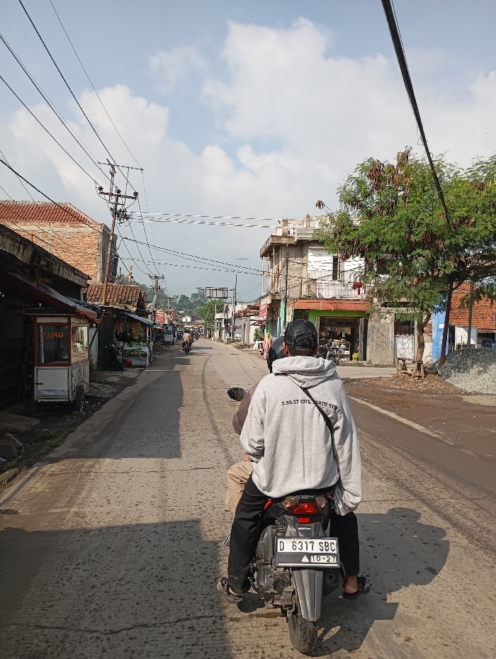 The image depicts a daytime street scene in Cipatik, Indonesia. A black Honda motorcycle with license plate D 6317 SBC is positioned in the foreground, traveling away from the viewer on a worn concrete or asphalt road. Two individuals are on the motorcycle: the driver, wearing a light grey hooded sweatshirt with the text "3.50.37 CITY NORTH STREET" on the back and a dark cap, and a passenger seated in front, wearing khaki pants.
The road is lined on both sides by a mix of single and multi-story commercial and residential buildings, some with red-tiled roofs or exposed concrete. On the left, a vendor stall with "TAHU" visible on a sign is present. On the right, a multi-story concrete building with balconies and a large tree with green and brown foliage are prominent. Numerous overhead electrical and communication wires crisscross the sky, supported by utility poles. Other motorcycles and figures are visible in the distance along the road. The background features distant hills or mountains under a partly cloudy blue sky.