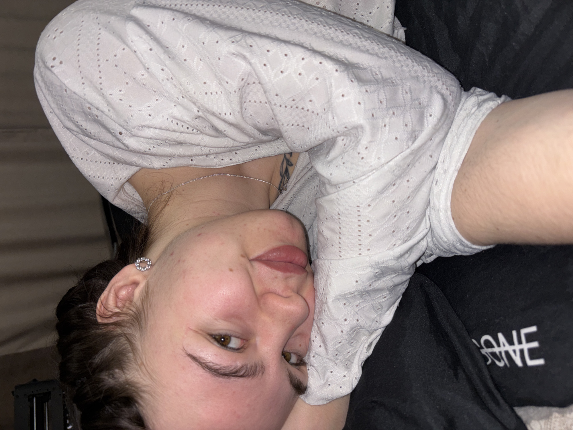 A woman with dark hair is lying down, wearing a white, patterned shirt and a silver necklace. She has a tattoo on her chest and a round earring in her left ear. She is partially resting on a black garment. The word "BONE" is visible on the garment. The photograph does not provide sufficient information to determine the location in Perm, Russia.