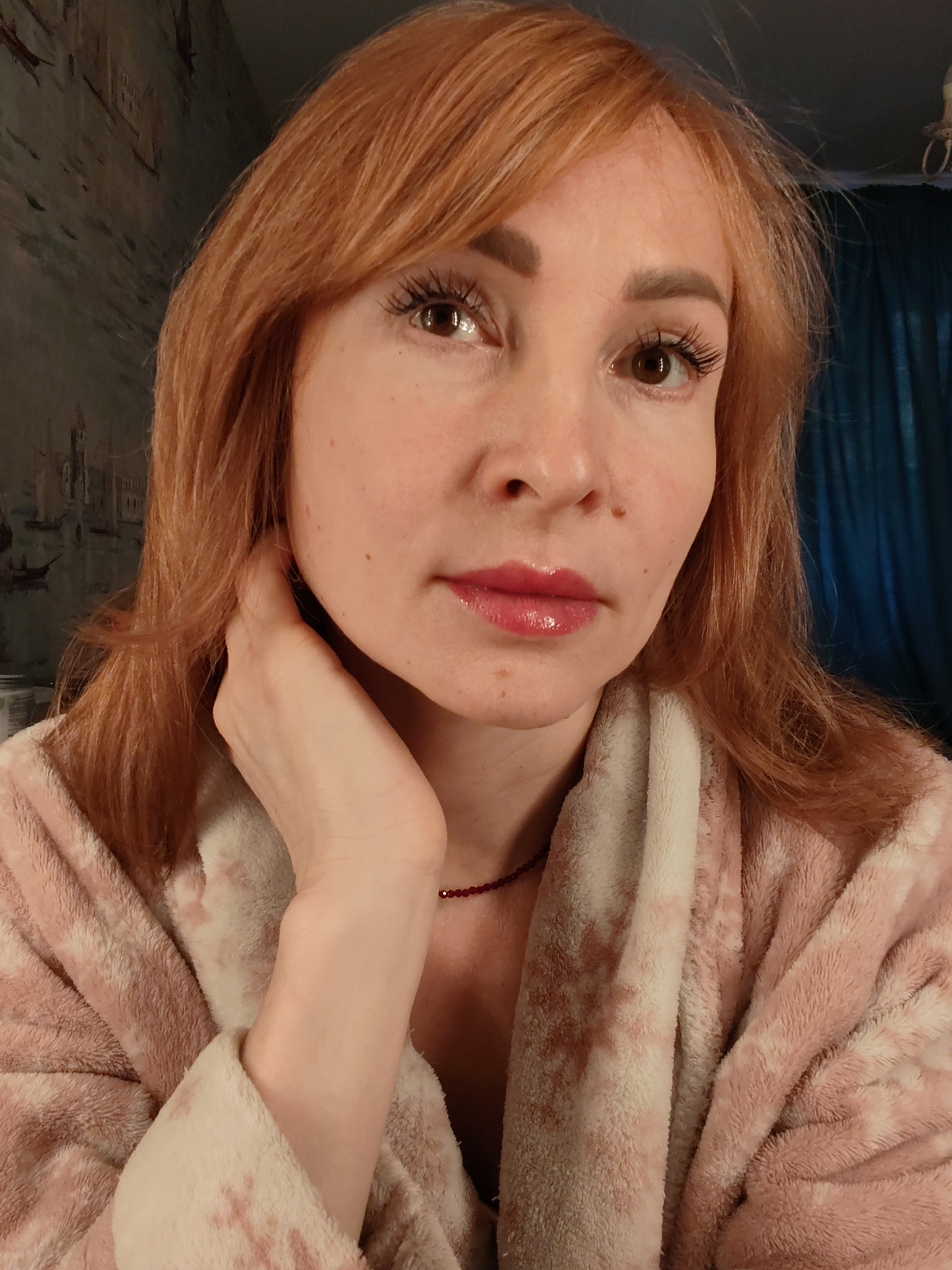 A woman with auburn hair, wearing a pink and white robe, is posing for a selfie. She has light skin, brown eyes, and is wearing lip gloss. Her hand is touching her neck. A dark red beaded necklace is visible. The background includes a textured wall with a faint pattern and a blue curtain. There is no indication of the location being Chaikovsky, Russia, or any other specific city.