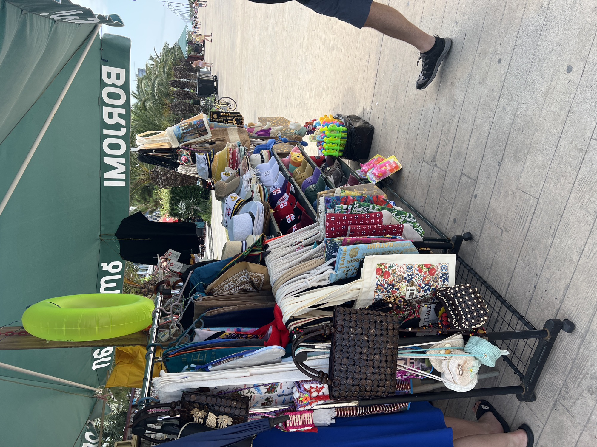 This image depicts a seaside promenade in Batumi, Georgia, likely near the beach area. A vendor stall displays an assortment of merchandise, including clothing, hats, bags, and beach accessories such as a bright green inflatable ring. The stall is situated under a green awning that features white lettering, partially obscured but appearing to read "BORJOMI." The promenade is paved with wooden planks, and palm trees line the background. Several people are visible in the distance walking along the promenade, enjoying the sunny weather. The overall scene suggests a tourist-oriented environment.