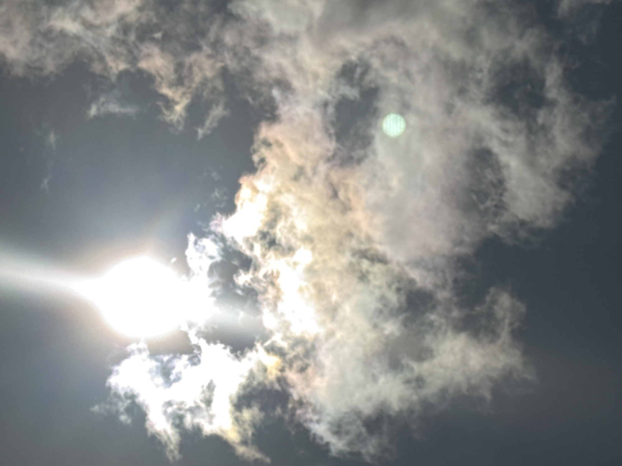The image depicts a cloudy sky with the sun shining brightly through. The sun is partially obscured by clouds, creating a halo effect and lens flare. Some of the clouds appear to have iridescent colors, possibly indicating a sun dog or other atmospheric optical phenomenon. There is a faint, circular, green object with a textured surface visible in the middle right portion of the image, which may be a lens artifact or a distant object. The sky is dark, suggesting it may be early morning or late evening, or that the clouds are thick. No specific landmarks or features are identifiable in the image to determine its location in Ivanovo, Russia. There are no people or discernible objects in the scene besides the sky and clouds.