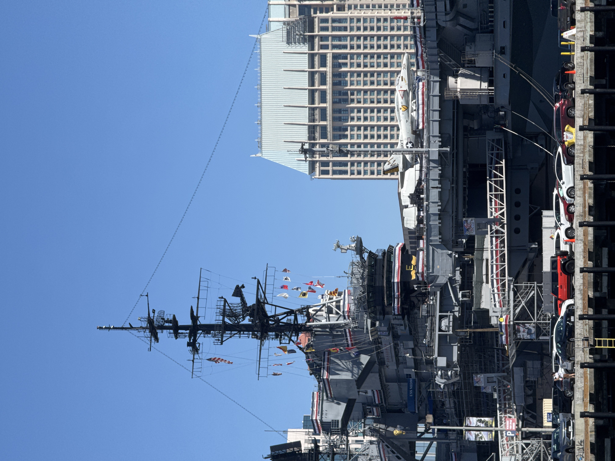 The image shows a view of the USS Midway Museum, an aircraft carrier museum located in San Diego, California. The aircraft carrier, identified by the text "USS MIDWAY" on its hull, is prominently featured in the foreground. It is docked at a pier with several cars parked along the waterfront. The ship has a tall mast with antennas and various flags flying. An airplane is displayed on the flight deck. In the background, a large, modern building with a distinctive light-colored, sloped roof stands against a clear blue sky. The scene depicts a sunny day in a harbor environment.