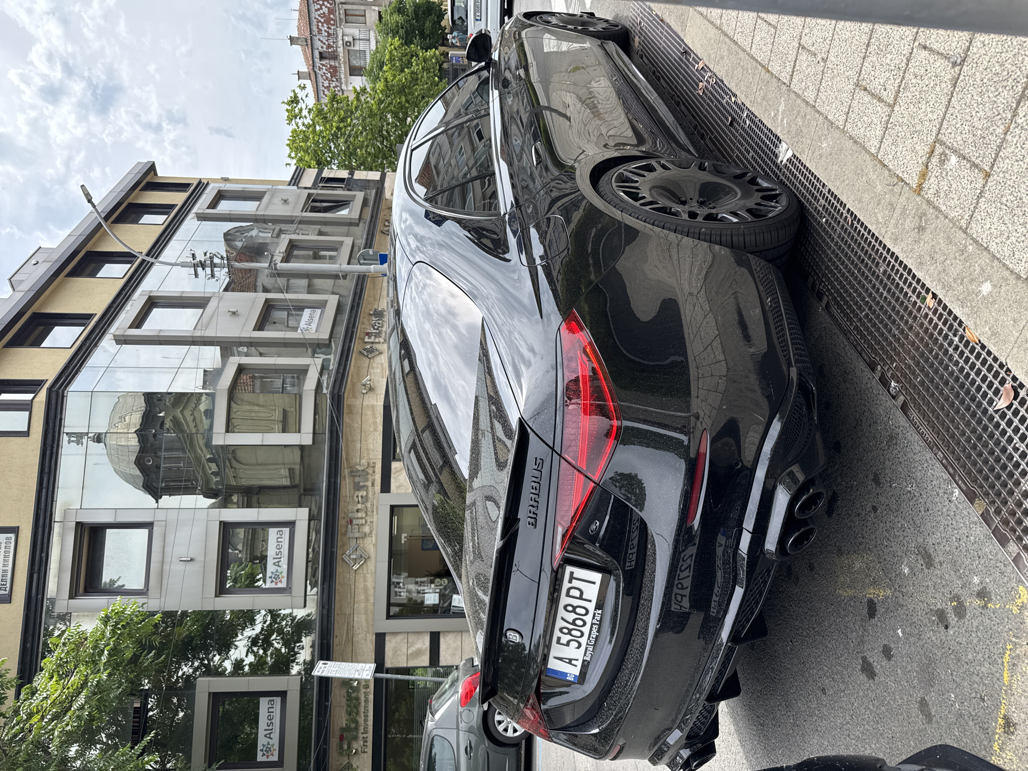 The image shows the rear three-quarters view of a black Brabus S-Class sedan parked on the side of a street in Burgas, Bulgaria. The car has a custom spoiler, quad exhaust pipes, and distinctive aftermarket wheels. The license plate reads "A 5868 PT". In the background, a modern building with a glass facade reflects the sky and an older building with a dome. The street is paved with cobblestones and has a metal grate drainage system along the curb. The scene appears to be in an urban setting during the daytime under an overcast sky. No people are clearly visible in the immediate vicinity of the car, though a blurred figure can be seen further down the street.