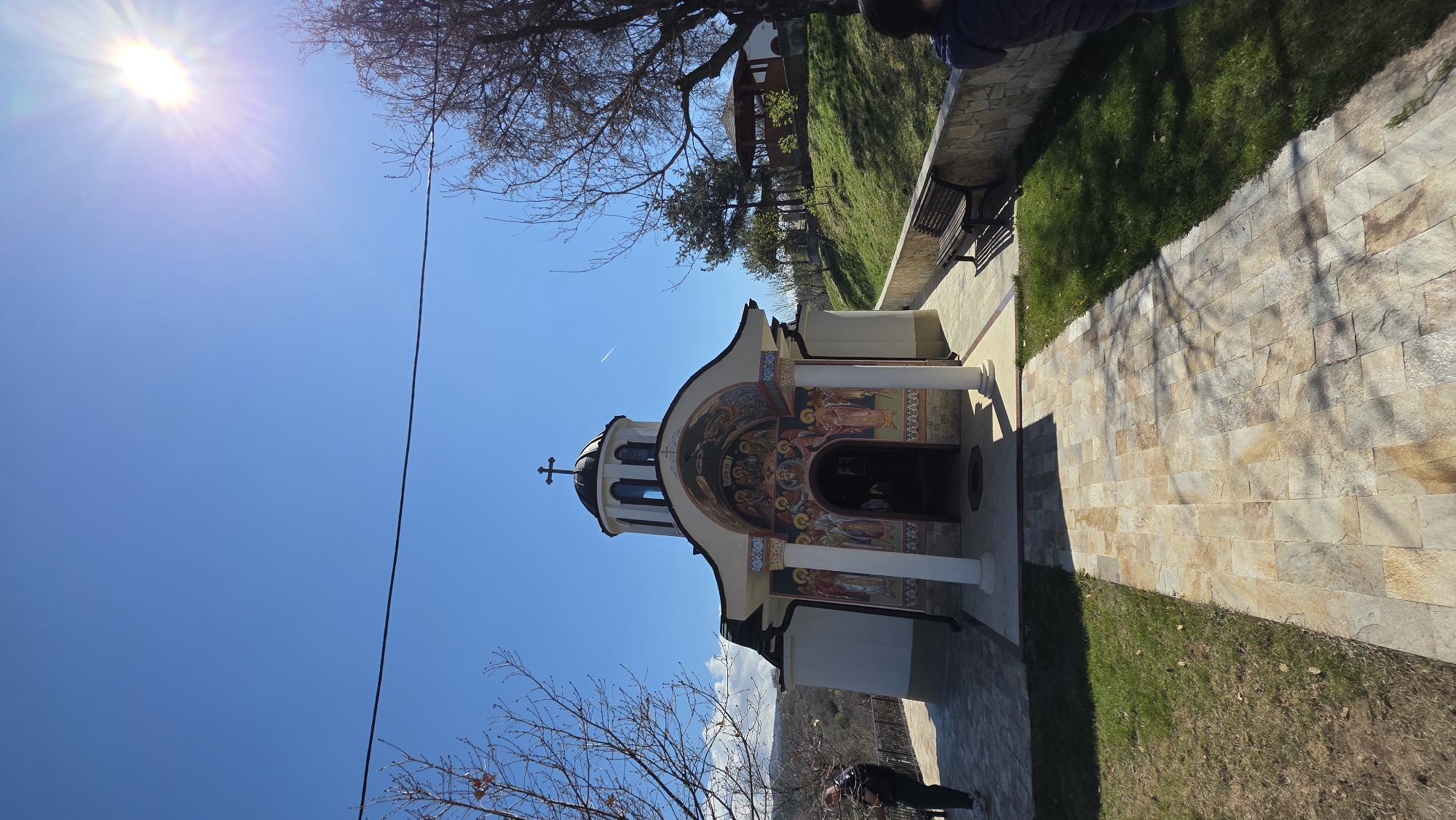 The image shows the St. Nicholas Church in Elenovo, Bulgaria, with the sun shining brightly from the upper left corner. The church is white with detailed painted murals around the entrance and a cross on top. A stone walkway leads to the church. To the right is a grassy area with a stone wall, and a wooden bench. There are trees in the foreground and background. A jet trail is visible in the sky. A person is visible in the bottom left and bottom right of the frame.