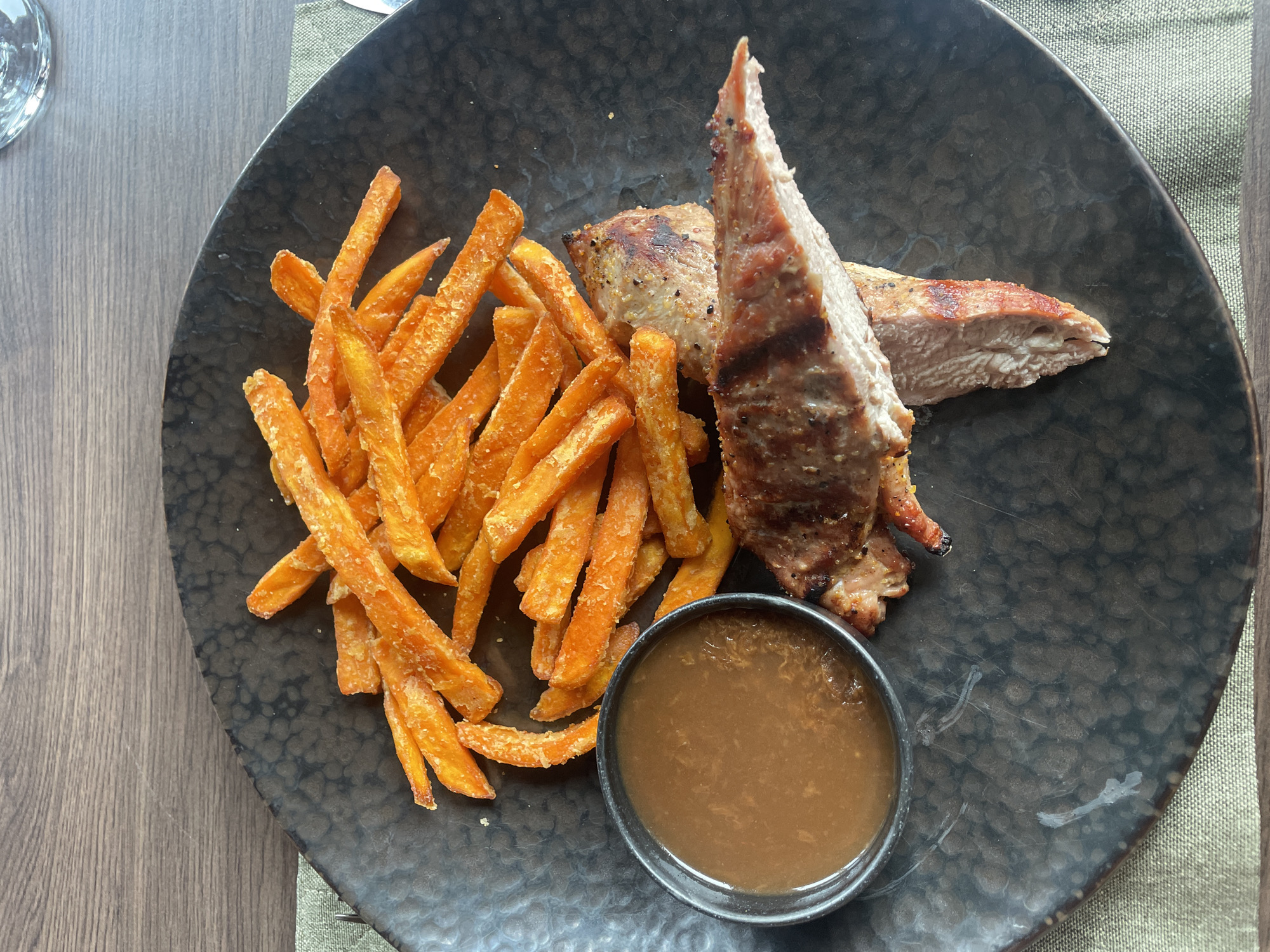 The image shows a plate of food. On the left side, there is a pile of sweet potato fries. In the center, there are two slices of grilled meat. There is a small bowl of brown sauce in the lower center. The plate is dark and speckled. The plate is set on a wooden table with a placemat. This may be a meal served in a restaurant. There is no information in the image that indicates a location in Pleven, Bulgaria.