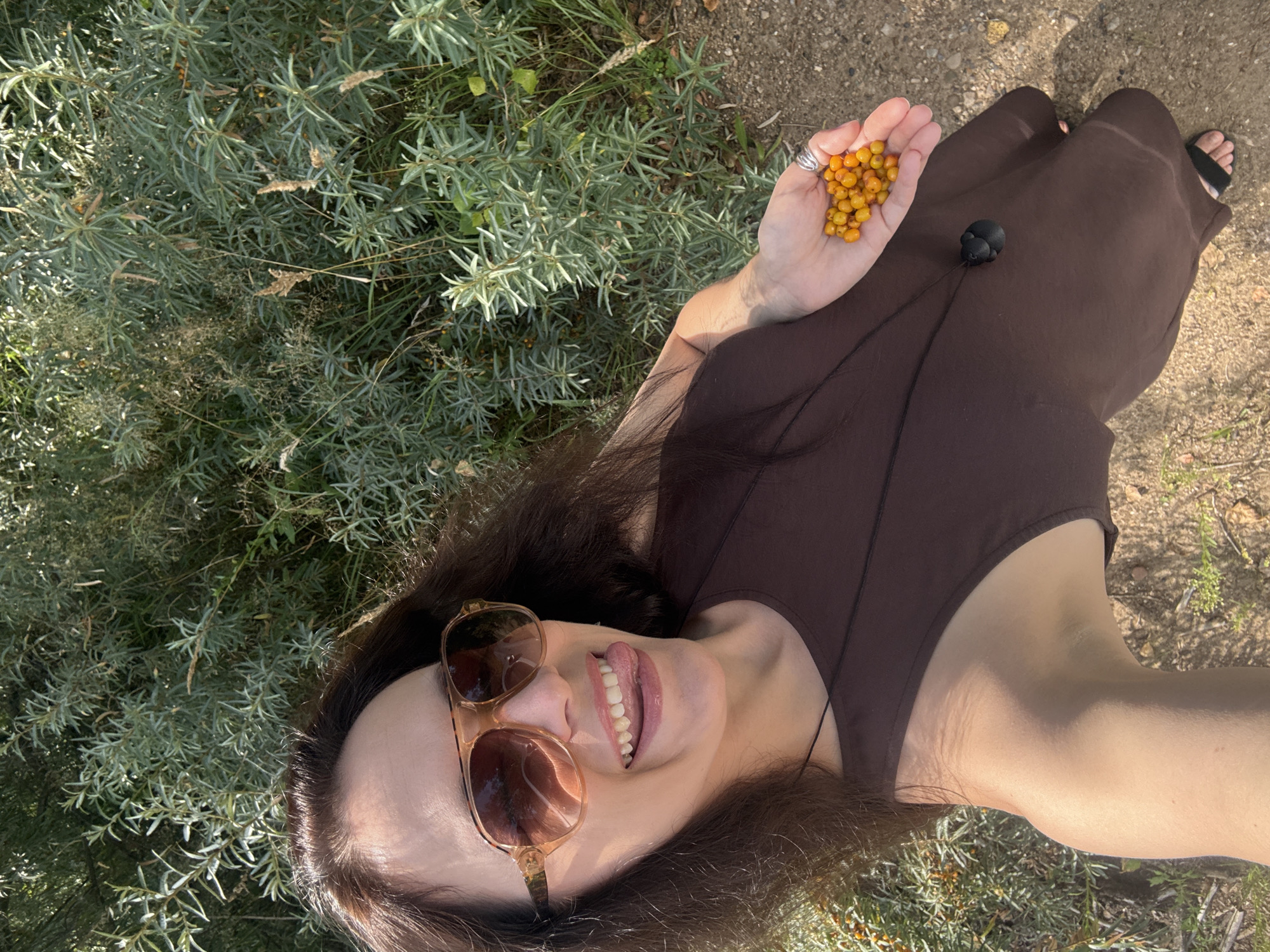 A woman is captured in a selfie outdoors, holding a small cluster of bright orange berries in her left palm. She is wearing a dark brown sleeveless dress and large, light brown aviator sunglasses. Her right hand is extended, offering a playful gesture with her tongue sticking out from between her teeth. The background consists of dense green foliage, likely bushes or trees, under a sunny sky. The ground beneath her is a mix of dirt and small stones. The overall impression is a candid shot of someone enjoying nature and possibly harvesting berries. The specific location in Chaikovsky, Russia, is suggested by the presence of sea buckthorn berries, which are common in the region and are known for their vibrant orange color and nutritional value.