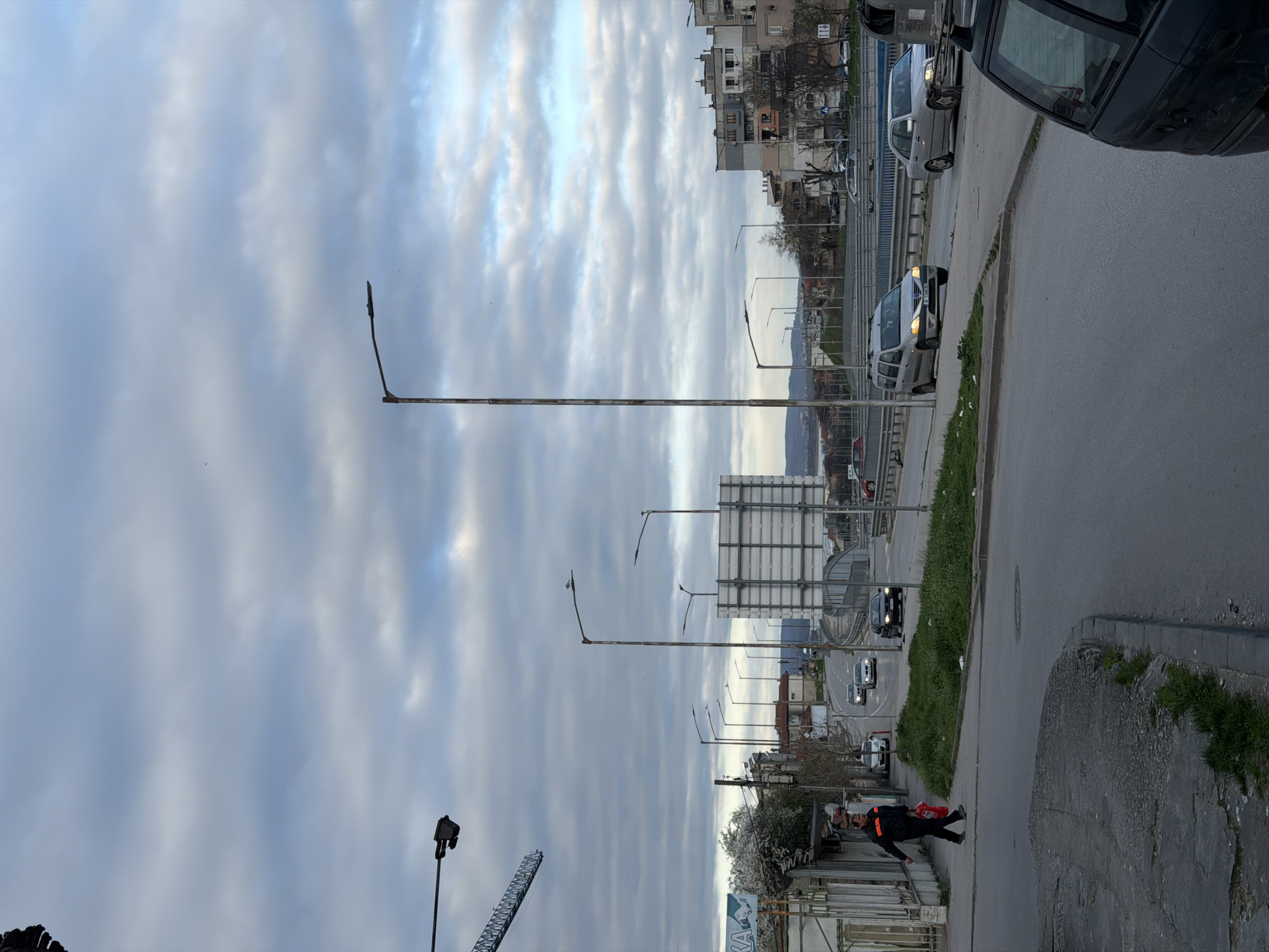 The image shows a street scene in Varna, Bulgaria. The view is facing a bridge and the street runs parallel to the bridge. There are streetlights along the street and a building in the foreground. A man wearing a black and orange outfit is walking on the sidewalk. Cars can be seen driving on the road. The sky is cloudy.