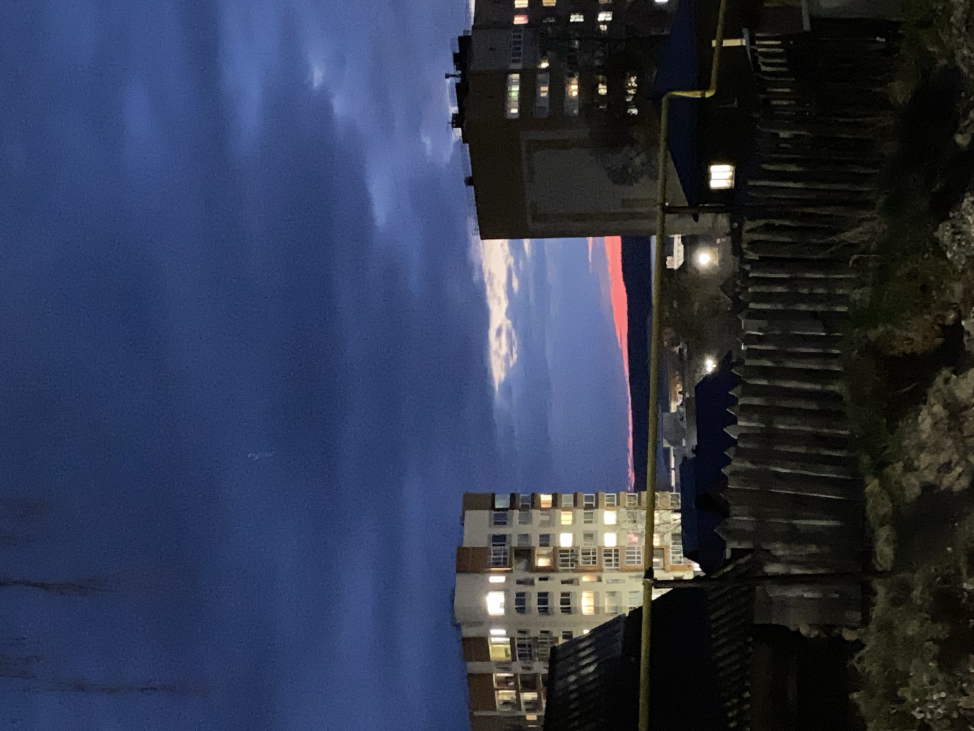 The image captures a nighttime scene in what appears to be an urban area of Votkinsk, Russia. Two high-rise apartment buildings dominate the view, with many windows lit up, suggesting residents are home. A deep blue, cloudy sky forms the backdrop, with a streak of pink possibly indicating the fading light of sunset. A dilapidated wooden structure and some vegetation are visible in the foreground, adding to the urban landscape. A yellow pipe is seen in the lower right side of the building in the bottom right. There are no people visible.