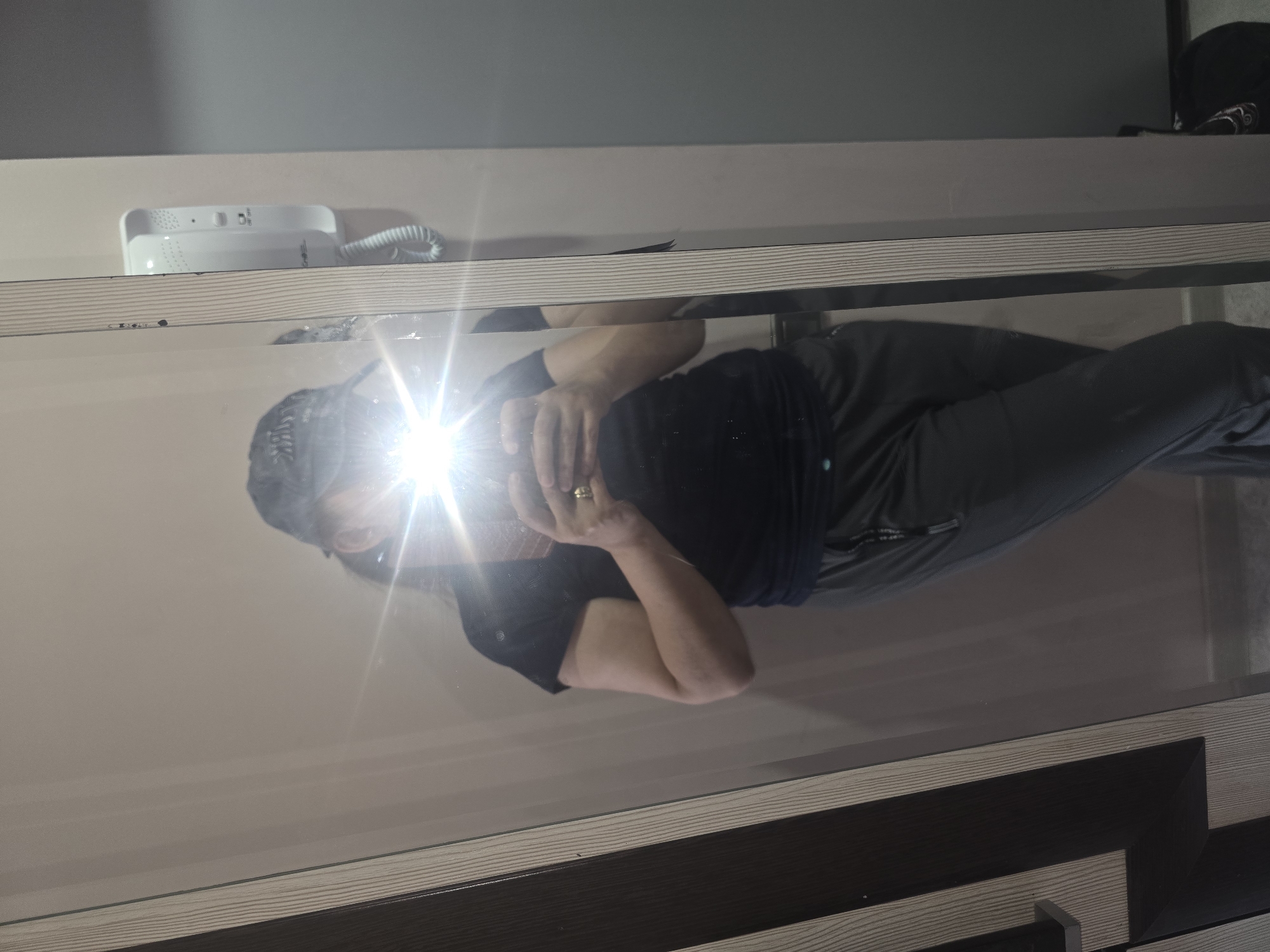 A person wearing a dark t-shirt and gray sweatpants, with a dark baseball cap, is reflected in a full-length mirror. The person is holding a phone to take a selfie, and a bright flash obscures the face. The mirror is set within a frame with vertical wood-grain detailing. To the right of the mirror, on the wall, is a white intercom device. The background consists of a wall and a portion of a dark gray door or cabinet on the far right. The scene appears to be indoors, possibly in a home or a hotel room. There is no visual information in the image to suggest the location is Ulan-Ude, Russia.