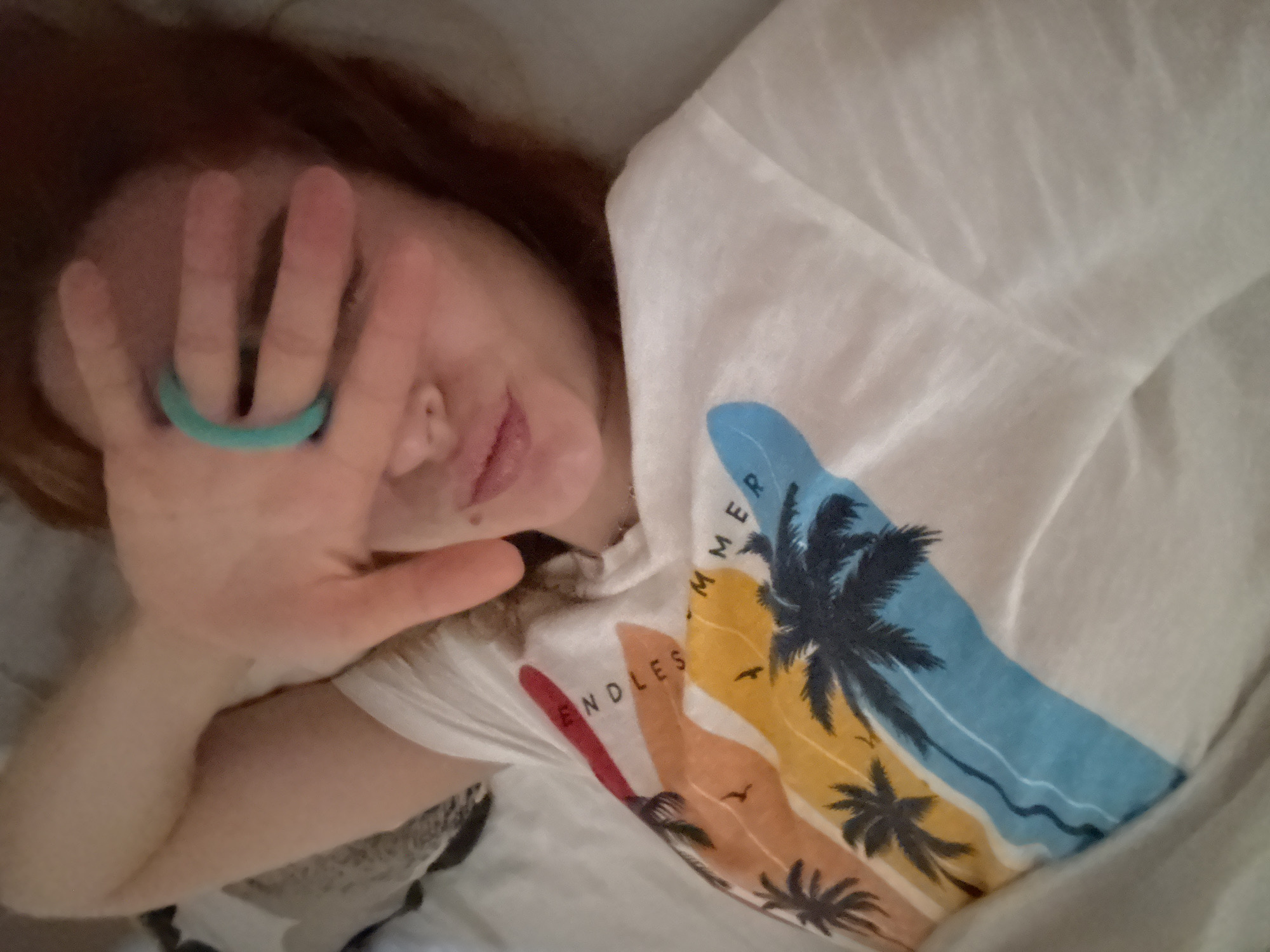 A woman with auburn hair lies on a bed, partially covered by a white sheet, wearing a white t-shirt with a colorful tropical design of palm trees and the words "Endless Summer." She holds her left hand up to her face, fingers splayed, with a turquoise ring on her ring finger. Her facial expression is partially visible, showing a slight smile. The location in Budva, Montenegro, is not discernible from the image.