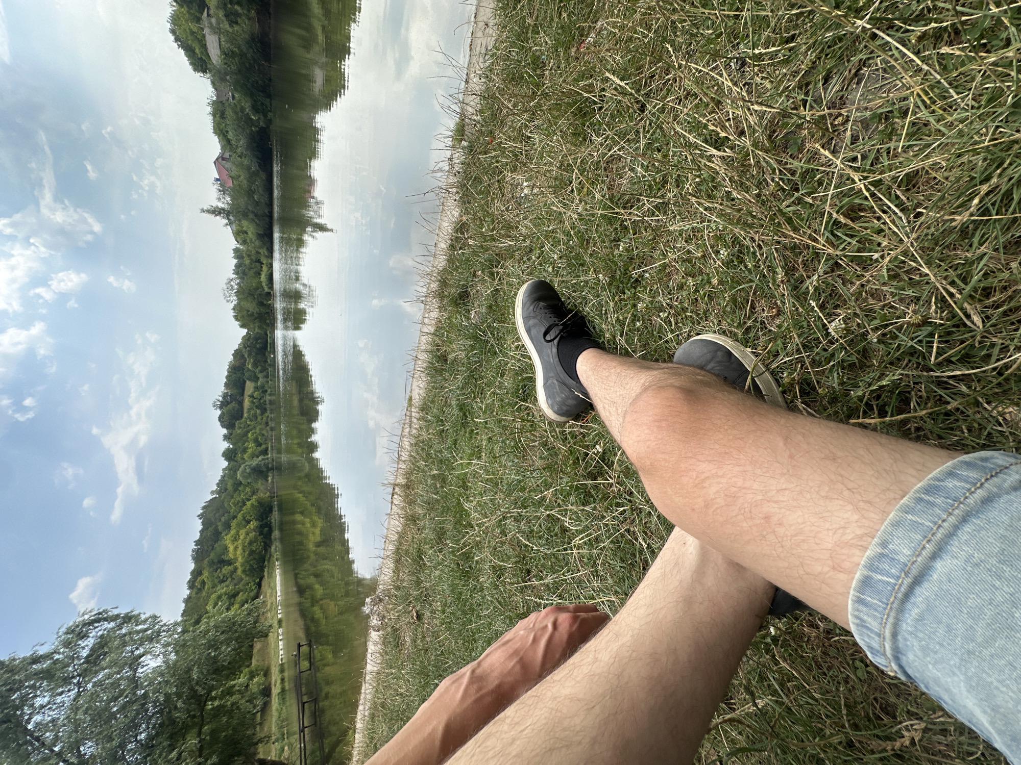 The image depicts a peaceful scene at a lake in Makiv, Ukraine. The foreground is dominated by the legs of a person sitting on a grassy bank. The person is wearing denim shorts and dark sneakers with black socks. Their right leg is bent at the knee, revealing tanned skin. The left leg is extended, and their hand rests on their thigh.
The background features a tranquil lake, its surface reflecting the cloudy sky and the lush green trees that line its shores. A small wooden pier is visible on the left side of the lake. In the distance, houses and more trees can be seen nestled amongst the hills. The overall atmosphere is serene and idyllic.