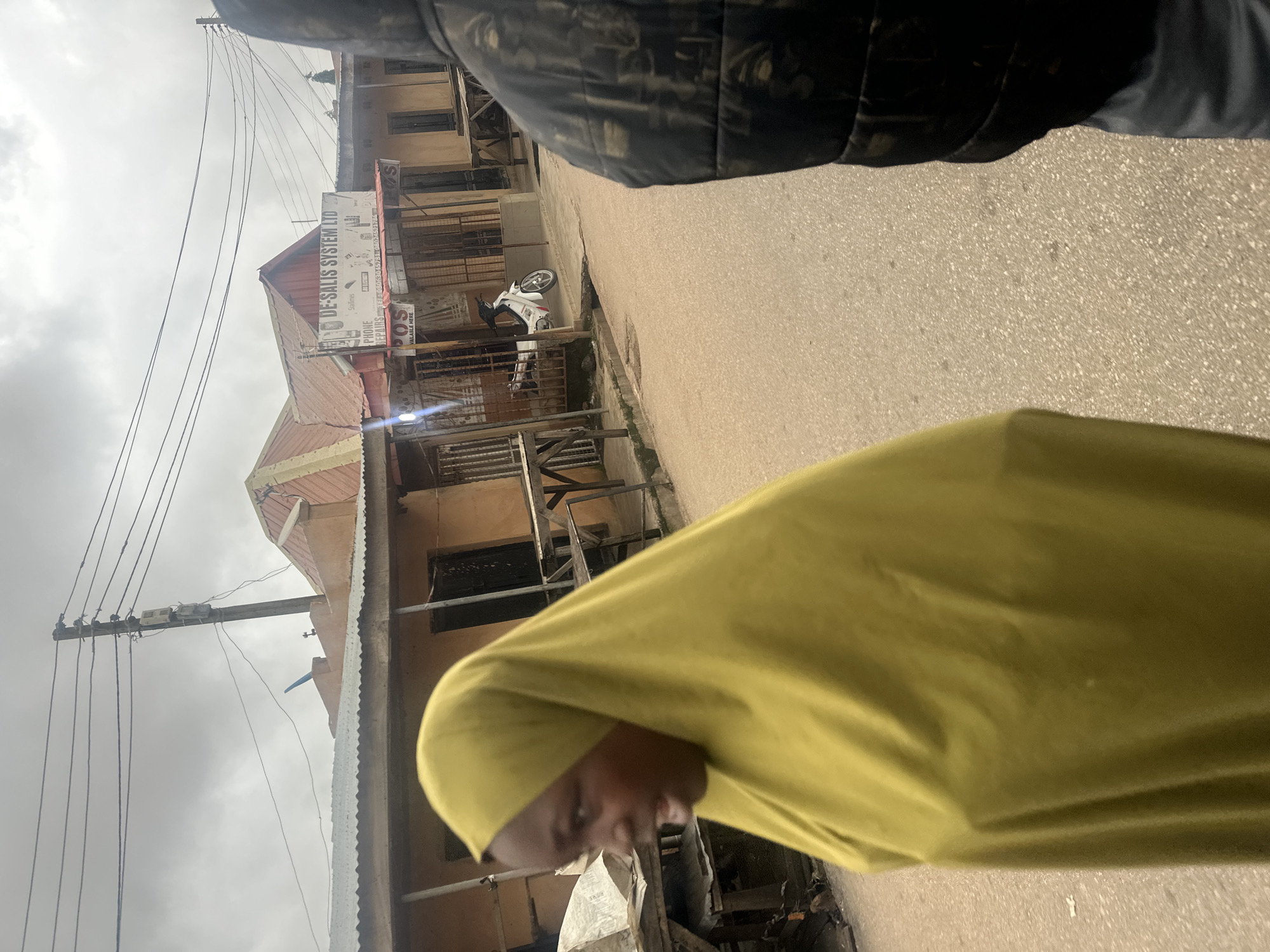The image depicts a street scene in Jos, Nigeria. In the foreground, a person wearing a bright yellow hijab is partially visible on the left, facing right with their mouth open as if speaking. To the right, another person, largely obscured from view, is wearing a dark, padded jacket.
The background features a row of shops with corrugated metal roofs. One shop has a sign that reads "DE-SALIS SYSTEM LTD" and advertises "PHONE REPAIRS." Below this sign, another smaller sign indicates "POS AVAILABLE HERE." A white motorcycle is parked in front of one of the shops. The street is paved with asphalt and shows signs of wear. Overhead, power lines are strung between utility poles, contributing to the urban landscape. The sky is overcast, suggesting a cloudy day. The overall impression is a glimpse into everyday commerce and street life in a Nigerian city.