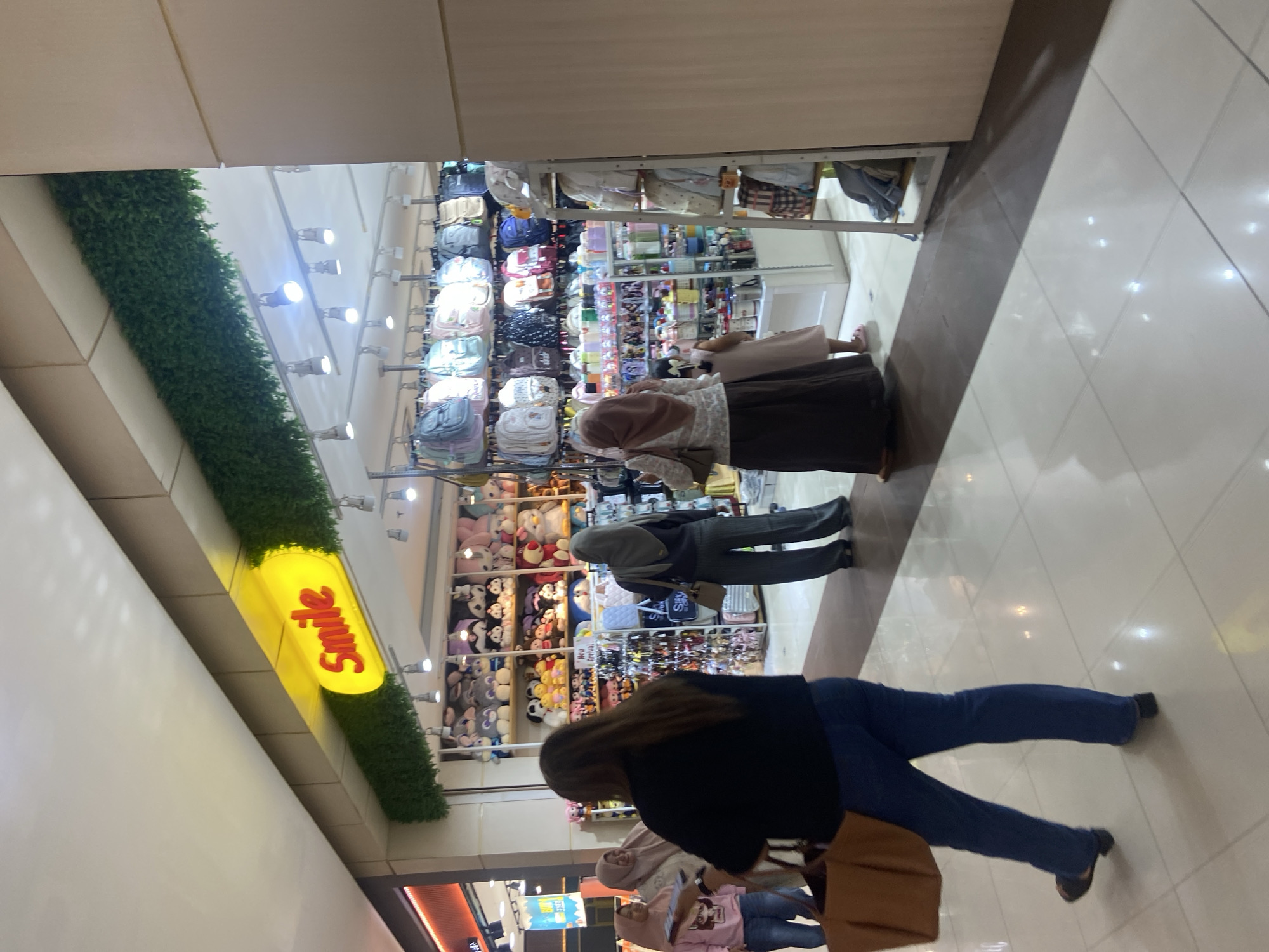In Sidoarjo, Indonesia, inside a brightly lit retail establishment identified by a prominent yellow sign reading "Smile," a variety of merchandise is displayed. The store appears to be a boutique specializing in accessories and plush toys, with shelves stocked with colorful backpacks, stuffed animals, and various smaller items. Several individuals are visible within the store. In the foreground, a woman with long brown hair, wearing a black top and blue jeans, walks away from the camera, carrying a large brown tote bag. To her left, two women wearing hijabs are browsing. One woman in a pink long-sleeved shirt looks at a phone, while the other, wearing a beige hijab, stands beside her. Further into the store, another woman in a grey hijab and striped pants is seen looking at merchandise. A young child in a light pink dress stands near the shelves. The flooring is a tiled surface, reflecting the ambient light. The store's interior is designed with a modern aesthetic, featuring clean lines and ample lighting.
