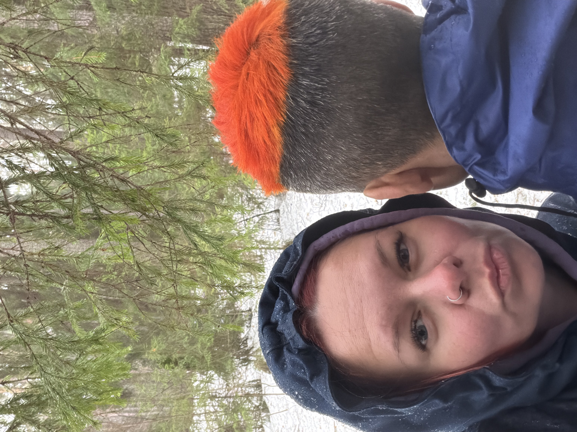 A selfie of two people in a snowy, wooded outdoor setting. A person with a gray and orange haircut and a blue jacket is positioned at the top of the frame. Another person with auburn hair partially visible under a navy blue hooded jacket with a purple lining is in the lower half of the frame, looking at the camera with a nose ring. Coniferous trees are visible in the background. Given the prompt, it's reasonable to assume this photo was taken in a wooded area near Chaikovsky, Russia.