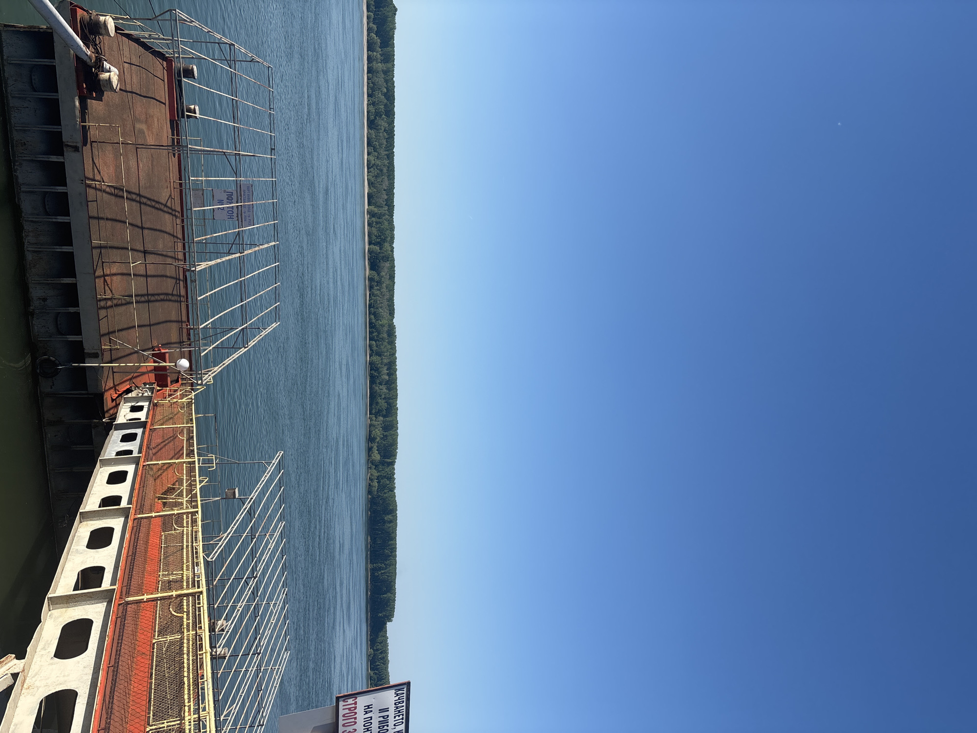 The image depicts a section of the Danube River in Lom, Bulgaria, viewed from the edge of a docked barge or ferry. The port infrastructure, comprising the barge’s metal framework, is visible on the left, with a red-painted deck and white supports. A line of trees marks the opposite riverbank. The right side of the image is dominated by a clear, blue sky.